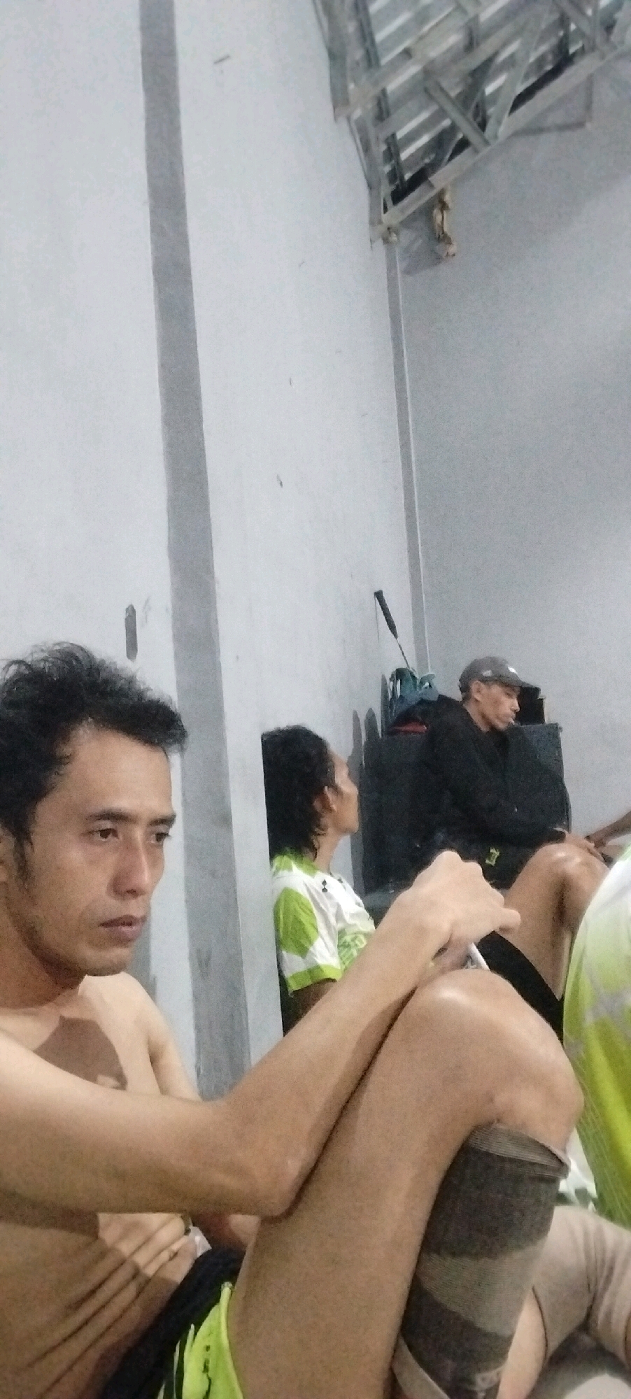 The image appears to be taken indoors, possibly in a recreational facility or community hall in Kliris, Indonesia. In the foreground, a shirtless man with short, dark hair is seated, looking to his left. He is wearing dark shorts with bright green accents. His right leg is bent at the knee, resting on his left leg, and a dark and grey band is visible around his right calf.
In the middle ground, another person is seated, facing away from the camera and looking to their right. This individual is wearing a white t-shirt with green patterned sleeves.
In the background, a man wearing a grey baseball cap and a black jacket is seated, looking to his right. He is positioned behind a large, dark object, possibly speakers or equipment. A black object, possibly a racket or a pole, is leaning against the wall behind him. The environment appears to be a simple, enclosed space with a plain wall and a metal ceiling structure. The lighting is dim, suggesting it might be evening or the interior is not brightly lit.