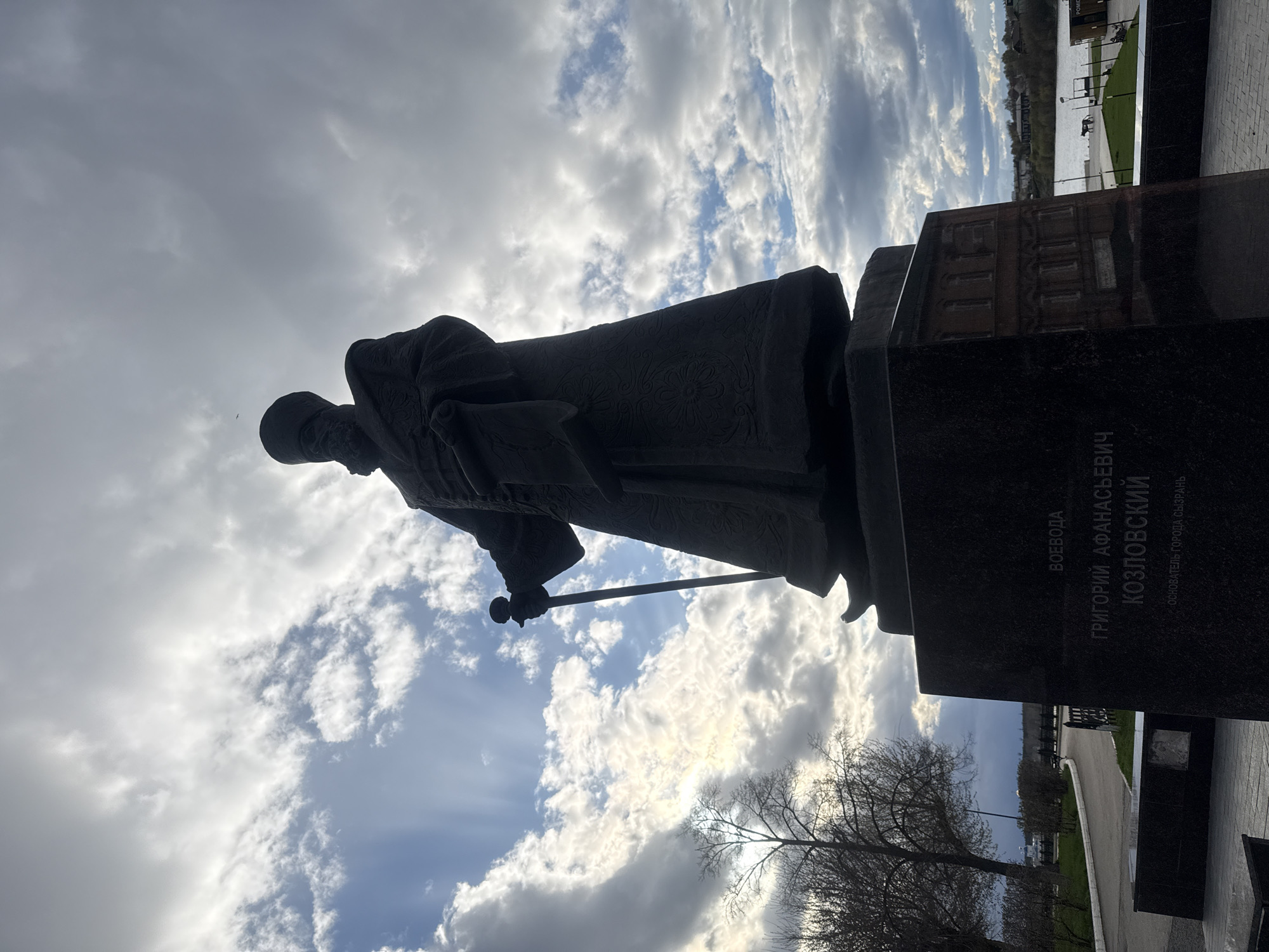 The image shows a statue of Grigory Afanasyevich Kozlovsky, a governor and the founder of the city of Syzran, Russia. The statue is made of a dark material, possibly bronze or a similar metal, and depicts Kozlovsky holding a staff. The statue is mounted on a dark stone pedestal, which bears an inscription in Russian. The background features a cloudy sky, a body of water with a park beside it, and some trees in the foreground.