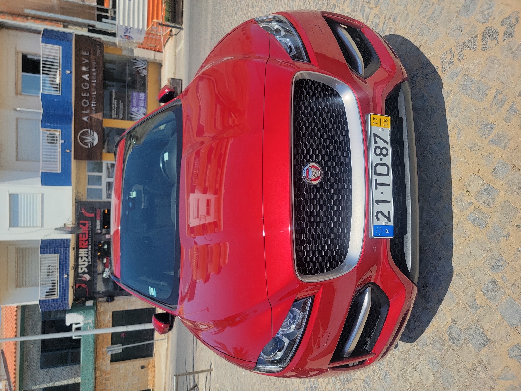 A front view of a shiny red Jaguar SUV is captured on a paved street in Quarteira, Portugal, on a sunny day. The car's license plate reads "21-TD-87 P". In the background, two commercial establishments are visible: "SUSHIREIKI" and "ALOEGARVE". The architecture features a blue and white facade, typical of Portuguese coastal towns. The street is made of cobblestones, and a shadow from the car is cast onto the ground.