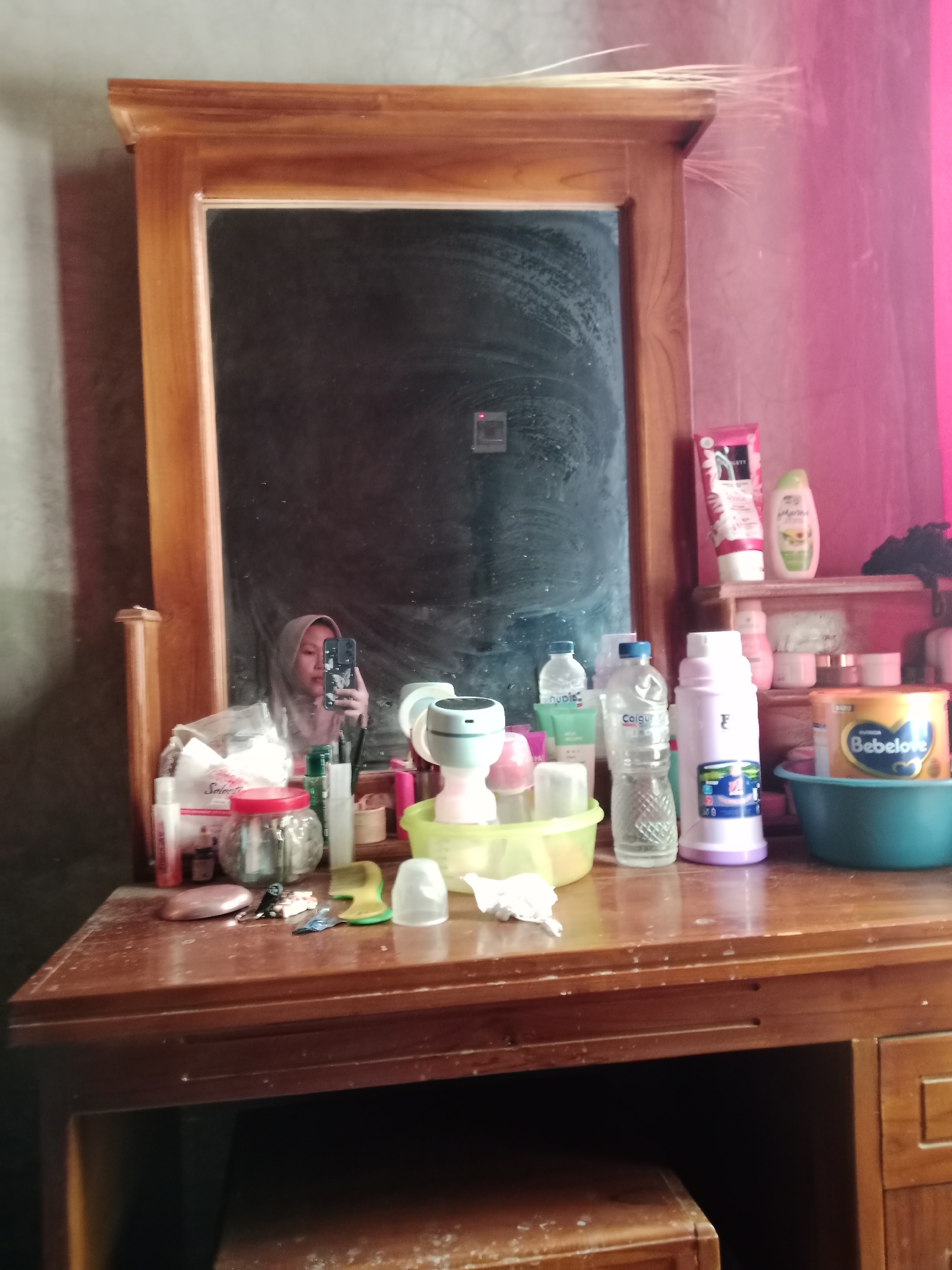 The image shows a wooden vanity table with a mirror. On the table are various toiletries, including bottles of lotion, shampoo, and a container of baby formula. A woman in a hijab is taking a selfie in the mirror. The room appears to be a bedroom. The city of Sindangjaya is located in Indonesia.