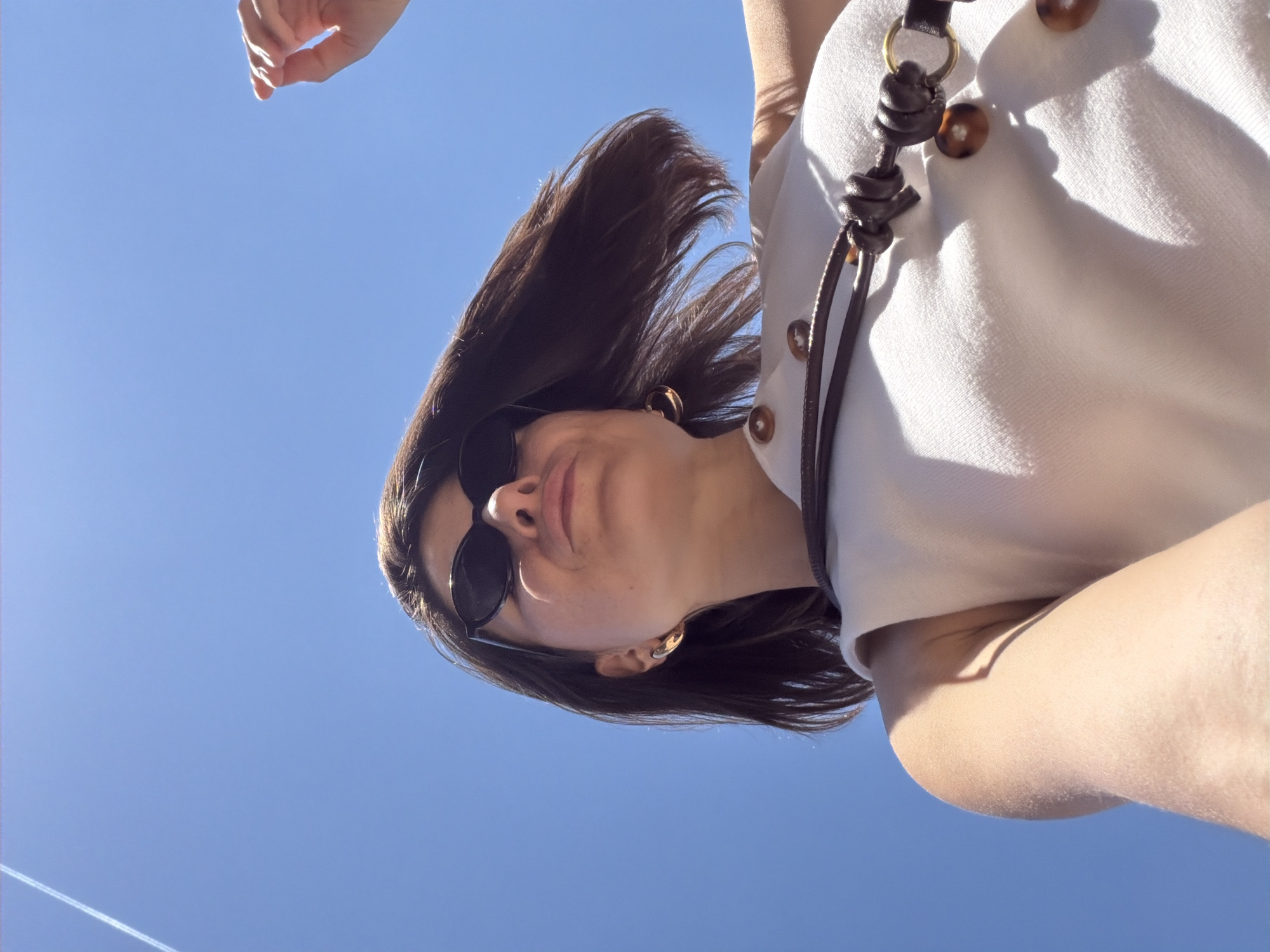 The image presents a low-angle, upside-down perspective of a person, likely female, against a clear blue sky. The subject's face is partially obscured by dark sunglasses, with her lips forming a slight pout. Her dark brown hair appears to be in motion, possibly wind-swept, and is illuminated by bright sunlight.
She is wearing a light-colored top, possibly white or off-white, featuring several round, light brown buttons down the front. A dark brown, braided leather-like strap with decorative knots and a brass-colored ring is visible on her shoulder, suggesting an accessory like a bag strap or glasses chain. A gold-toned earring is visible on her right ear. A hand, reaching upwards, is partially visible at the top of the frame.
The background is a vibrant, cloudless blue sky. A faint, straight white contrail from an aircraft is visible in the lower left portion of the sky. The strong lighting indicates bright sunshine and daytime, casting defined shadows on the person's clothing and skin, particularly under the chin and around the face. The overall mood is casual and natural, with the unusual angle adding dynamism. The scene is set in Budva, Montenegro. There is no visible text.