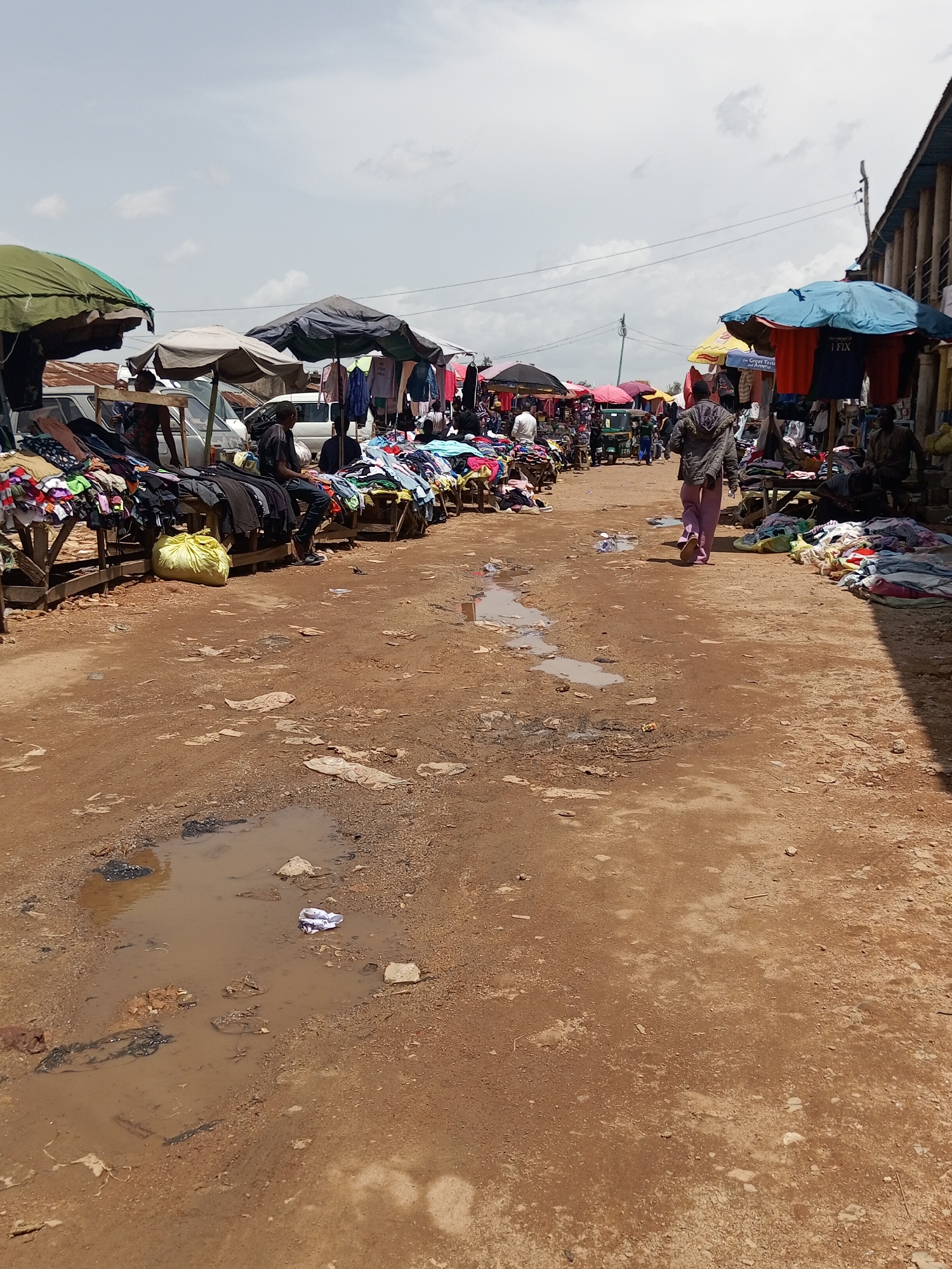 The image depicts a bustling outdoor market scene in Bukuru, Nigeria. The ground is a dirt path, uneven and marked by several puddles of muddy water, indicating recent rainfall or poor drainage. Stalls line both sides of the path, covered by various colorful umbrellas and awnings, predominantly in shades of green, blue, and red. These stalls are laden with a wide assortment of clothing, neatly folded and displayed on wooden tables or hung on racks.
Several individuals are present, engaged in the activity of buying and selling. In the foreground, a person walks away from the camera, wearing a patterned hooded jacket and pink trousers. On the left side of the market, a man in a dark shirt sits on a wooden bench, with clothing displayed in front of him. Other figures are visible in the background, some attending to stalls and others browsing. A small, three-wheeled vehicle, likely a tuk-tuk, is parked further down the path.
The sky is overcast with scattered clouds, suggesting it might be daytime with potentially humid or post-rain weather. The overall impression is one of a vibrant local marketplace, typical of many in Nigeria. One stall prominently displays a blue umbrella with red and white lettering; the visible text on a red garment hanging from it reads "I FIX". Another stall has a yellow umbrella with text that includes "Great Taste". The scene is alive with activity, conveying a sense of commerce and community.