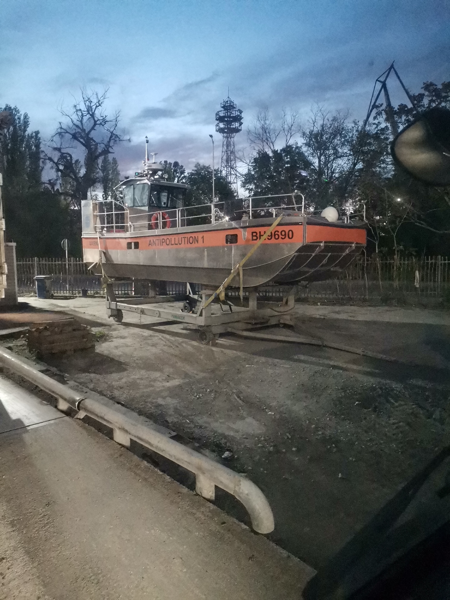 The image depicts an "Antipollution 1" vessel bearing the registration number BH.9690, situated on a trailer within an industrial area. The vessel is silver and orange. The area appears to be located in Varna, Bulgaria as "VARNA" is written on the side of the boat. The environment consists of concrete or paved surfaces interspersed with a rougher, gravelly area. A metal guardrail runs alongside the paved surface, and fencing is visible in the background. The lighting suggests it is dusk or night. A metal tower and bare trees are in the background.