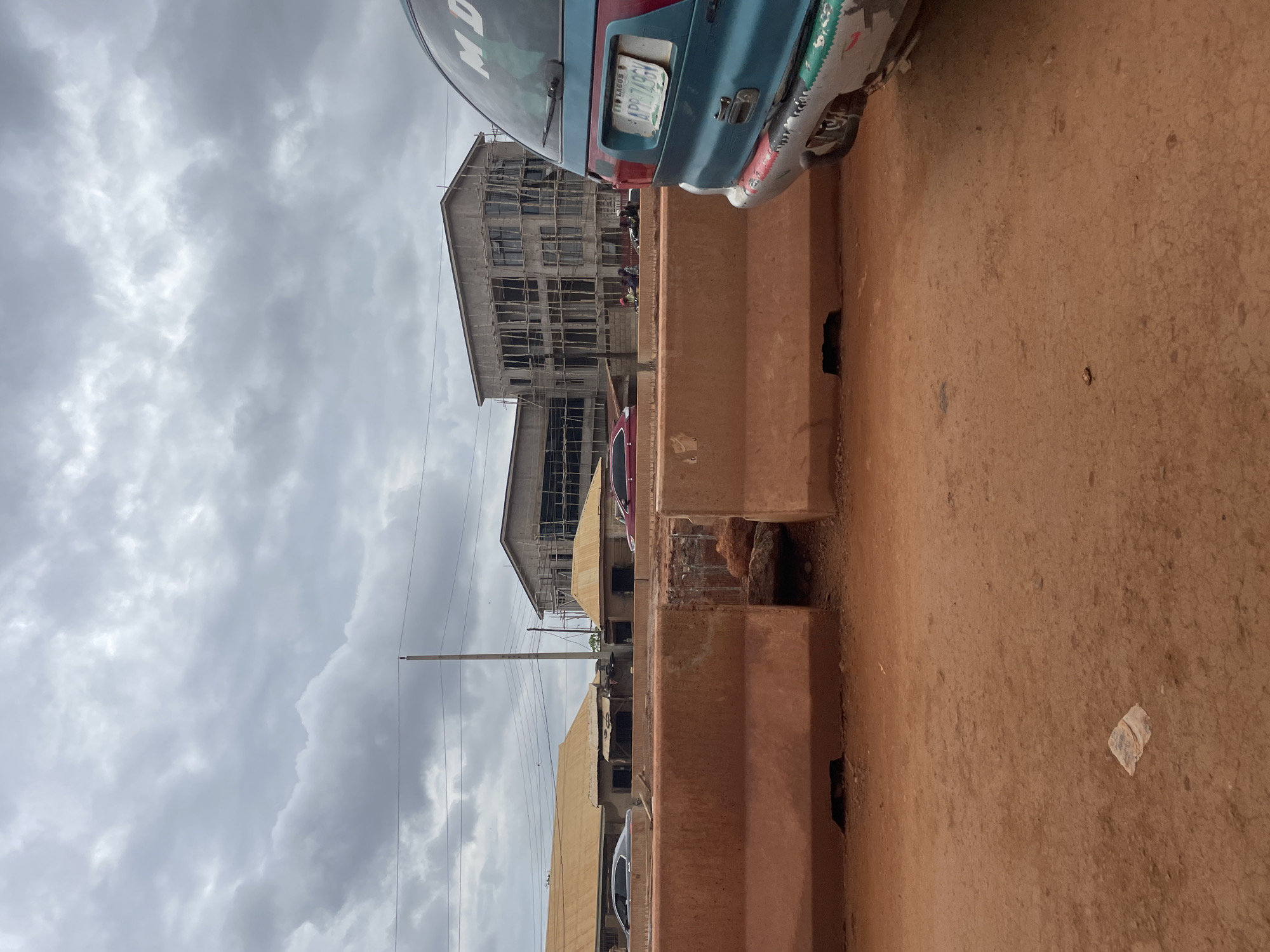 A cloudy sky looms over a construction site in Minna, Nigeria. In the foreground, orange concrete barriers separate a dirt road from the ongoing construction. A turquoise minivan with a Lagos license plate is parked on the right side of the frame, its rear facing the viewer. Behind the barriers, several buildings are under construction, with scaffolding visible on one multi-story structure. Various vehicles, including a silver sedan and a red car, are present on a road running parallel to the construction site. Small structures with yellowish roofs are also visible, suggesting an area with mixed development. The ground is covered in reddish-brown dirt, with some scattered debris.