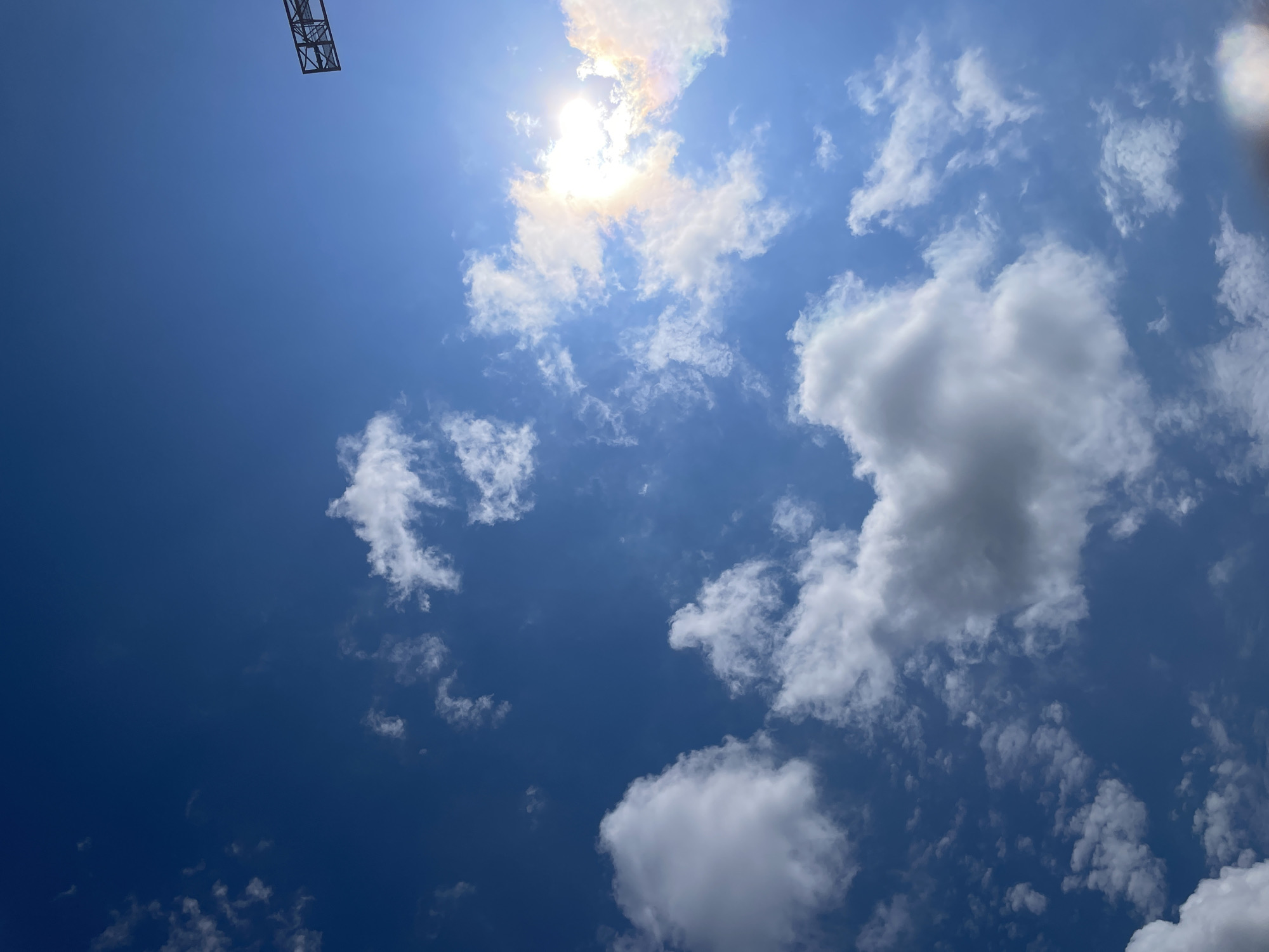 The image displays a bright blue sky filled with scattered white clouds. The sun is shining intensely, creating a lens flare and a faint rainbow effect on the right side of the image. In the upper right corner, a portion of a dark, metallic structure, possibly a signpost or part of a stadium, is visible against the sky. The perspective is looking upwards, emphasizing the vastness of the sky. No people or specific landmarks of Stavropol, Russia, are identifiable in this image.