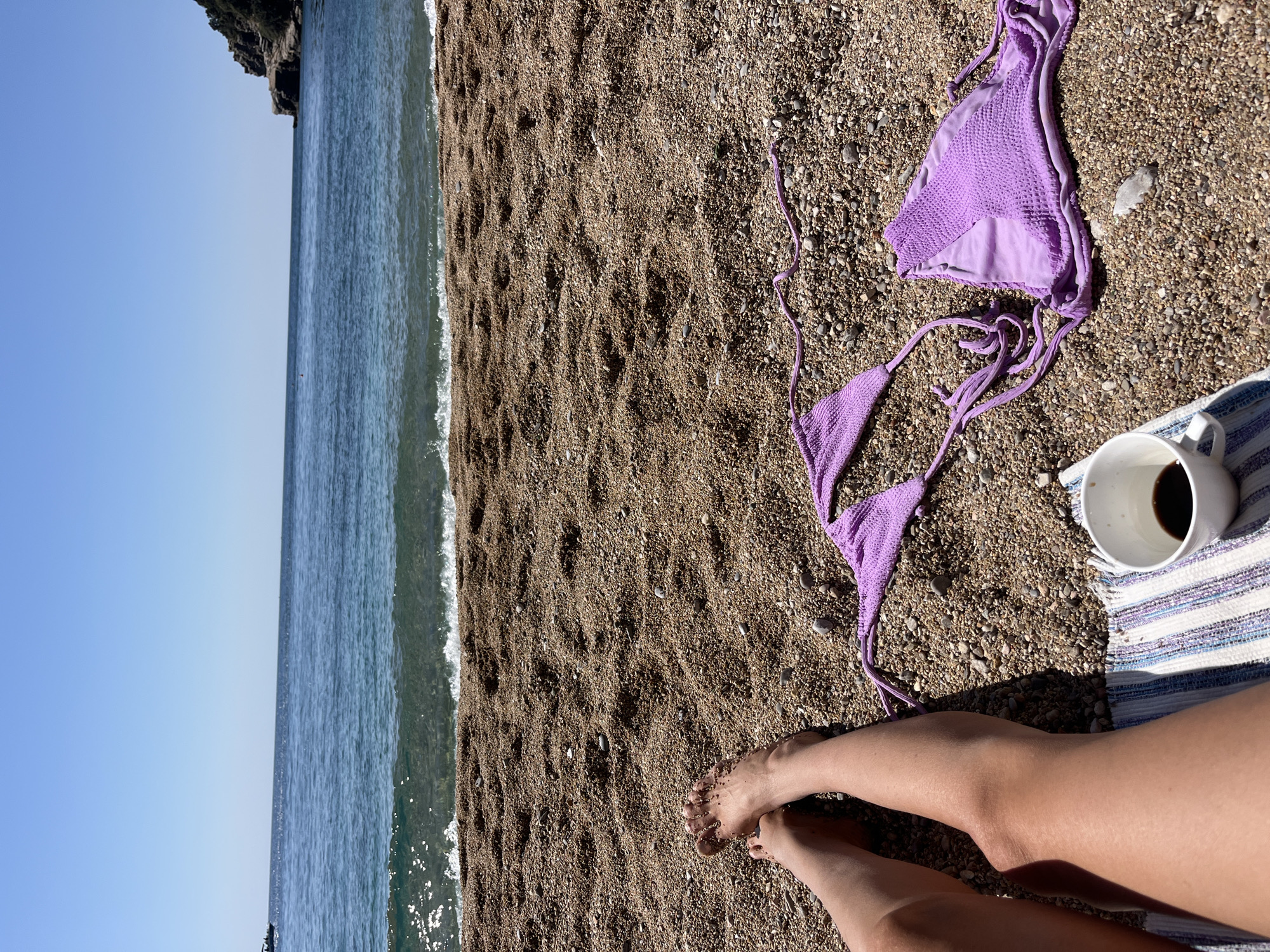 The image depicts a beach scene in Budva, Montenegro. A shoreline with a calm, teal-colored sea meets a pebble beach. The sky is clear blue. A bikini top, light purple and textured, lies on the pebbles. A white mug filled with a dark liquid sits on a striped, blue and white towel. Bare legs are visible in the foreground, belonging to a person relaxing on the beach.