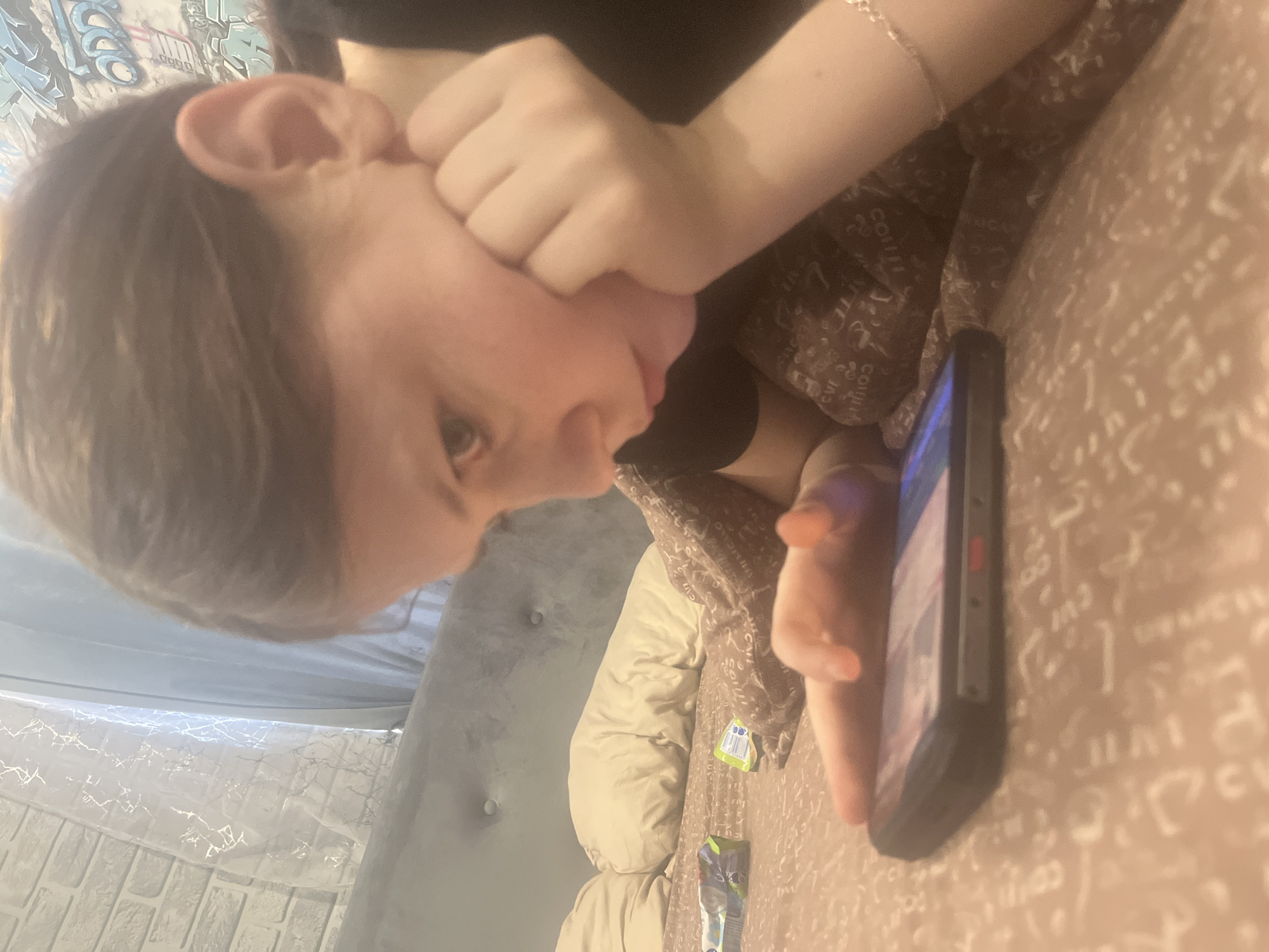 A young person is lying down on a couch while looking at a cell phone. The individual is propped up with a hand to the face and appears to be watching something on the screen. There is a blanket covering the couch, with various textures and patterns visible. A partial view of a headboard and wall are also in the frame. The image contains no information to indicate a location in Vesyoloye, Russia.