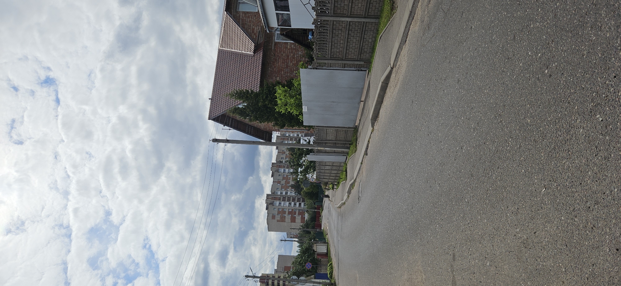The image depicts a street scene in Vitsebsk, Belarus. A paved road runs from the foreground into the distance, curving slightly to the left. On the right side of the road, there is a concrete fence with decorative elements, followed by a large, plain metal gate. Behind the fence, a two-story brick house with a brown tiled roof is visible, partially obscured by trees. To the left of the road, there is a sidewalk and further down the street, several multi-story apartment buildings can be seen. The sky is overcast with clouds. There are no people or vehicles visible in the image. A speed limit sign indicating 40 km/h is present on the left side of the road. Power lines are strung across the scene, supported by utility poles.