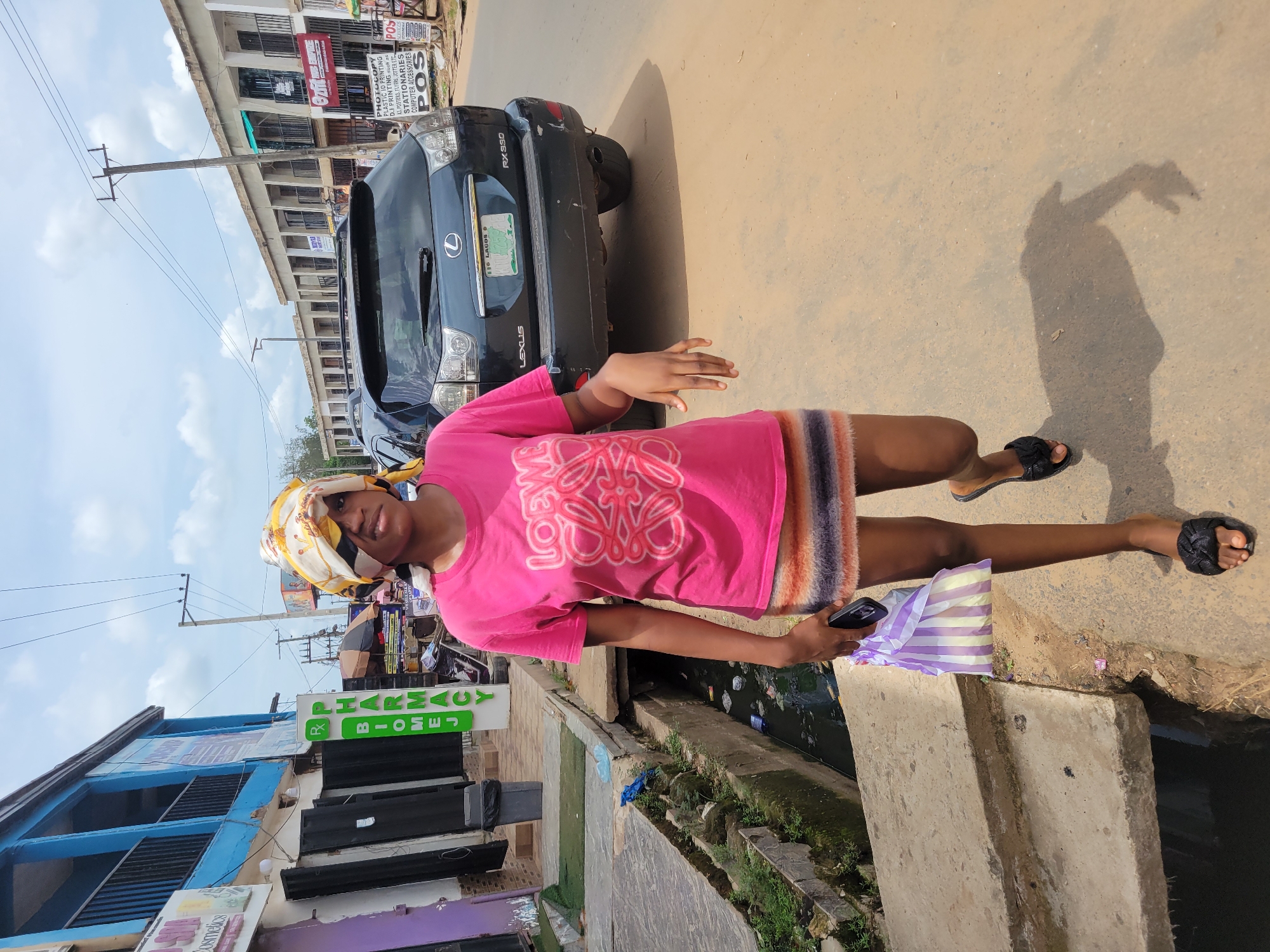A woman in a pink shirt and headscarf stands on a city street in Egbagu, Nigeria. A parked black Lexus RX 330 is visible in the background. Buildings with storefronts are across the street. A concrete structure, possibly a drainage canal, is next to the woman. A shopping bag is in her hand, and she is holding a mobile phone. Overhead power lines stretch across the sky.