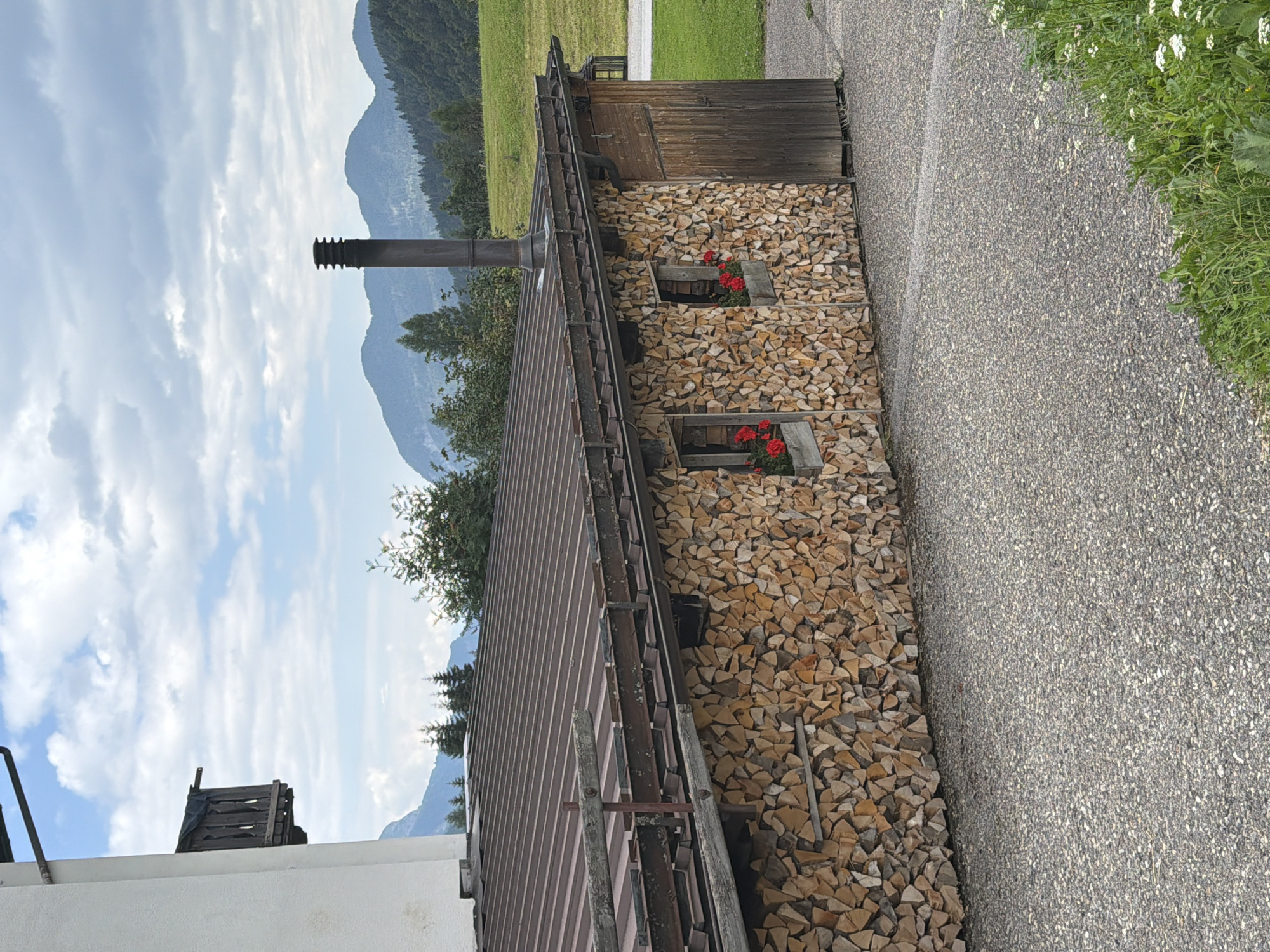 The image shows a rustic building with a wall made of stacked firewood. The building has a corrugated metal roof and a chimney. There are two small windows on the firewood wall, each with a planter box containing red flowers. A wooden door is visible on the right side of the building. The building is situated in a mountainous region with a paved driveway in the foreground. In the background, there are green hills and distant mountains under a cloudy sky. The scene appears to be in Sauris di Sopra / Plozn / Sauris Disore, Italy, given the architectural style and landscape.