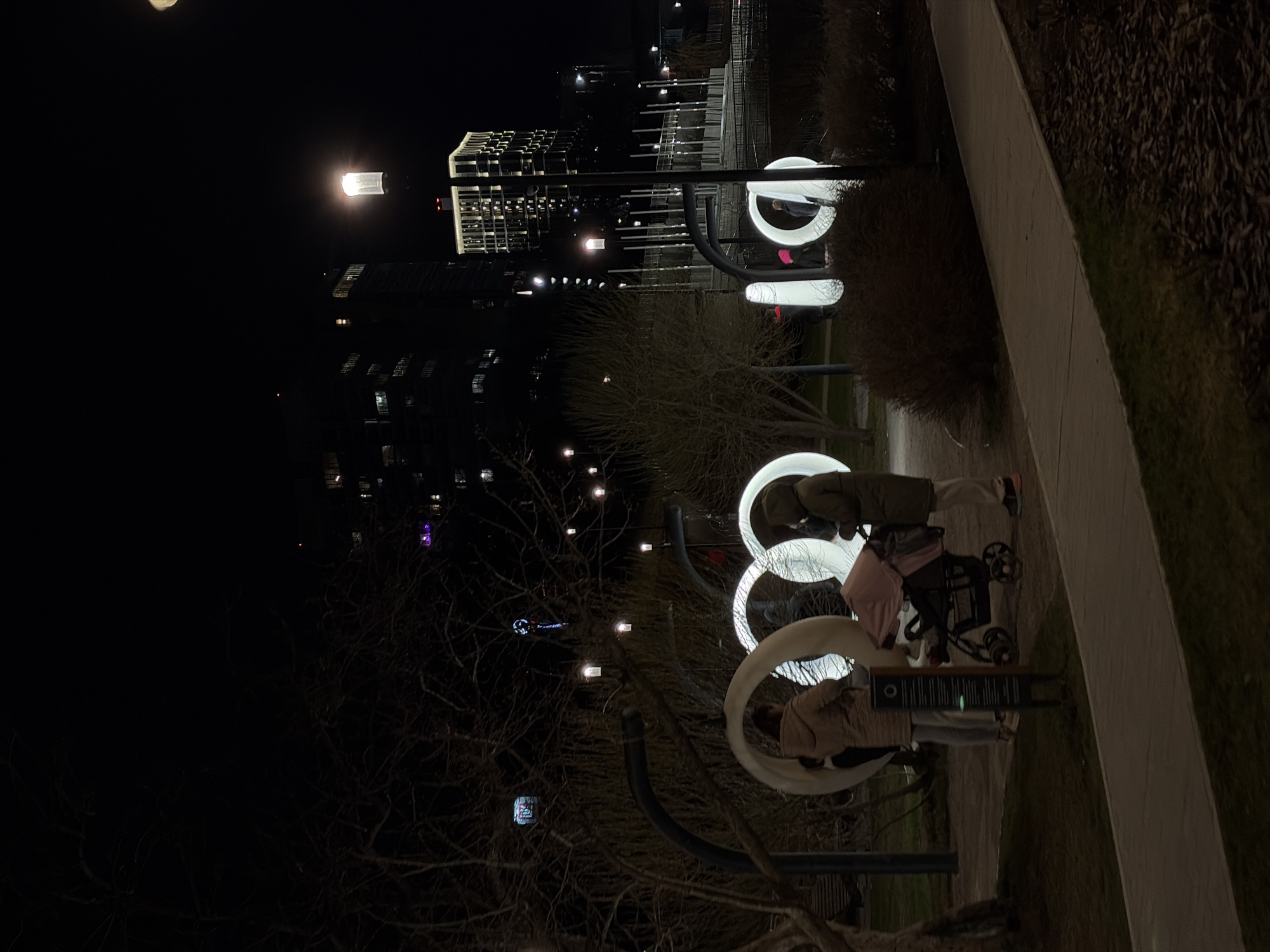The image captures an outdoor night scene, likely in a park or urban plaza in Perm, Russia. The primary focus is a series of large, white, circular or ring-shaped structures that are brightly illuminated, serving as striking art installations or unique seating.
Two adults and a baby in a stroller are present. One person, wearing a light-colored jacket and light pants, stands beside a stroller with a pink canopy, near one of the illuminated rings. Another person, in a darker jacket, is partially visible further back among the installations. They appear to be enjoying an evening walk or resting in the illuminated space.
A paved pathway stretches from the right foreground, bordered by dark grass and sparse bushes. Bare tree branches are visible on the left, suggesting a season without leaves. In the background, modern city buildings with numerous illuminated windows rise against the dark night sky. Several streetlights also contribute to the artificial illumination of the scene.
A dark, rectangular informational sign or panel stands near the person with the stroller. It features faint white text and a circular emblem, though the specific content is illegible due to distance and lighting. The overall atmosphere is tranquil, with the bright art installations contrasting sharply with the dark surroundings.