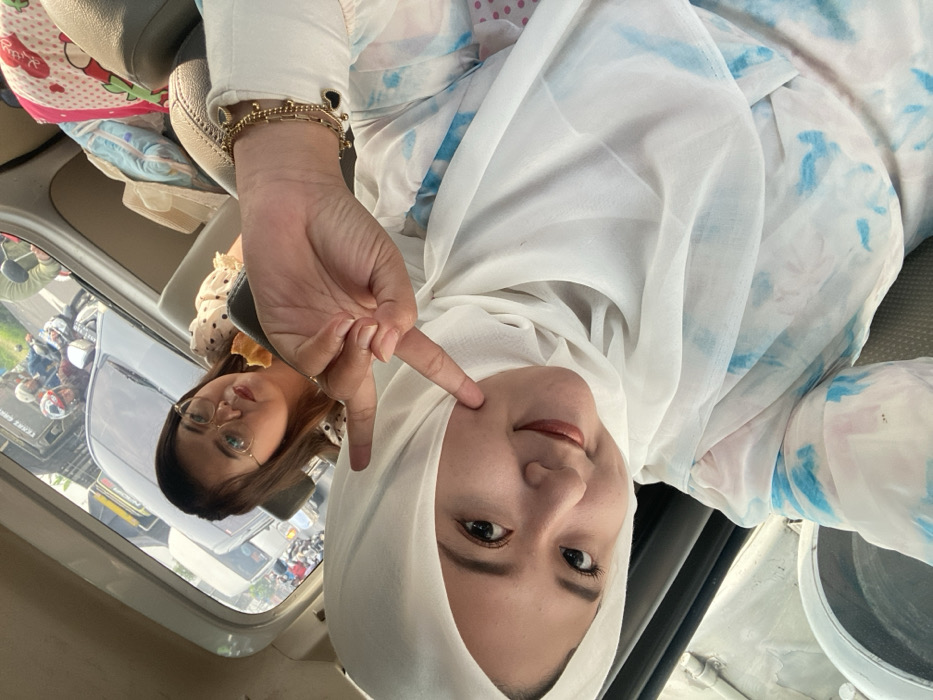 Two women pose inside a vehicle. One woman is wearing a white hijab and making a peace sign near her cheek. Her outfit is a white blouse with light blue patterns. She has a bracelet on her left wrist. In the reflection of the window, another woman is seen wearing glasses and a white polka dot blouse. The outside view shows traffic with motorbikes and other vehicles.