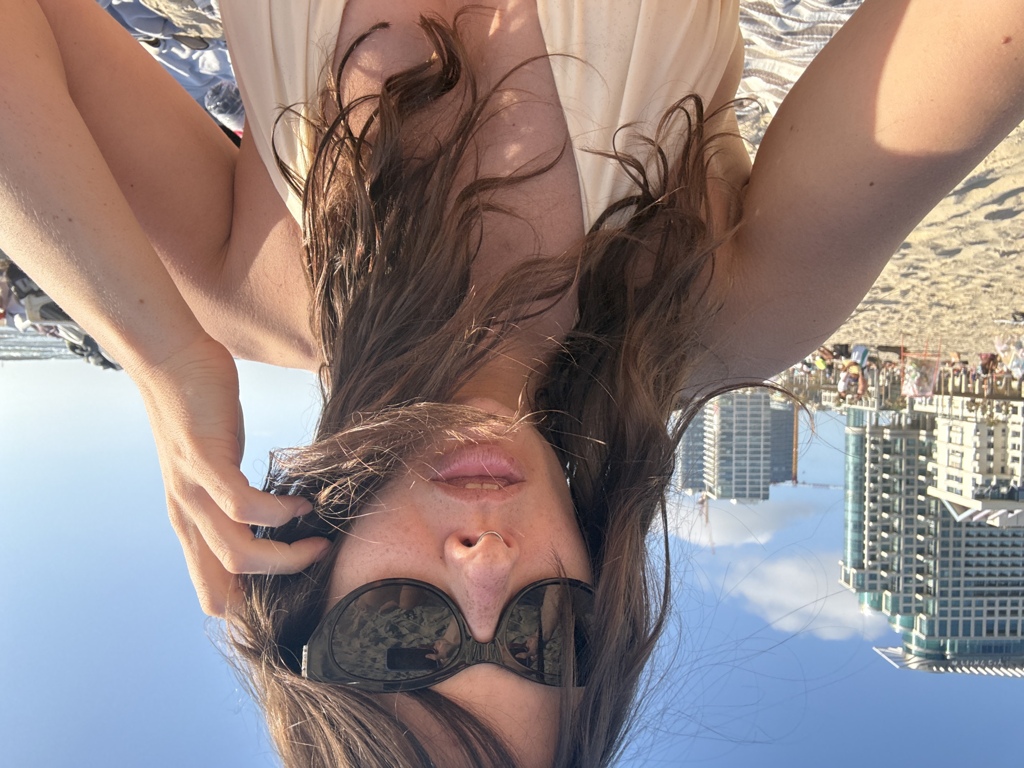 A selfie photograph taken by a person on a beach. The person is wearing sunglasses and is facing downwards towards the camera. Her hair is brown and partially covers her face. Her arms are raised on either side of her head. The background shows a beach scene with numerous people, a blue sky, and several tall buildings. These buildings are identifiable as part of the Tel Aviv skyline, indicating the location is likely on one of the beaches of Tel Aviv, Israel.