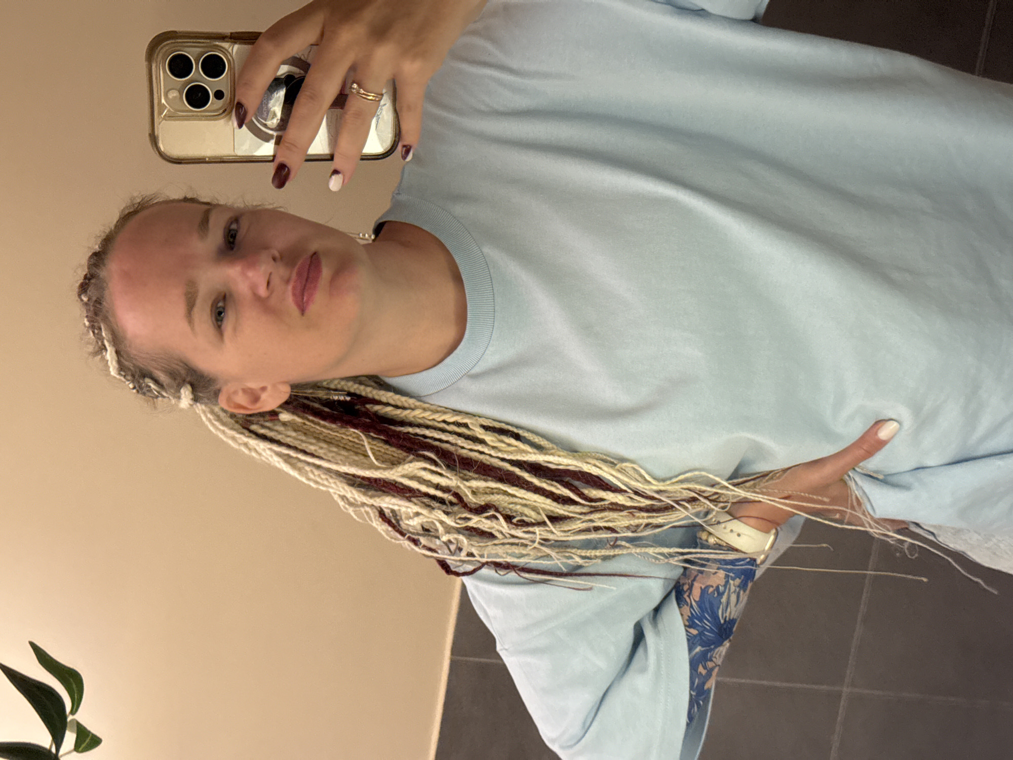 A person is taking a selfie in what appears to be a bathroom. The person has light skin and is wearing a light blue long-sleeved shirt. Their hair is styled in blonde and burgundy braids, some of which are draped over their shoulder. The person is wearing a gold ring on their right ring finger and a white watch on their left wrist. In the reflection of the phone, their fingernails are painted with a dark color on one hand and white and dark on the other. The background is a neutral-colored wall, and a dark tiled floor is visible. A plant with dark green leaves is partially visible in the lower left corner. The lighting suggests it could be daytime.