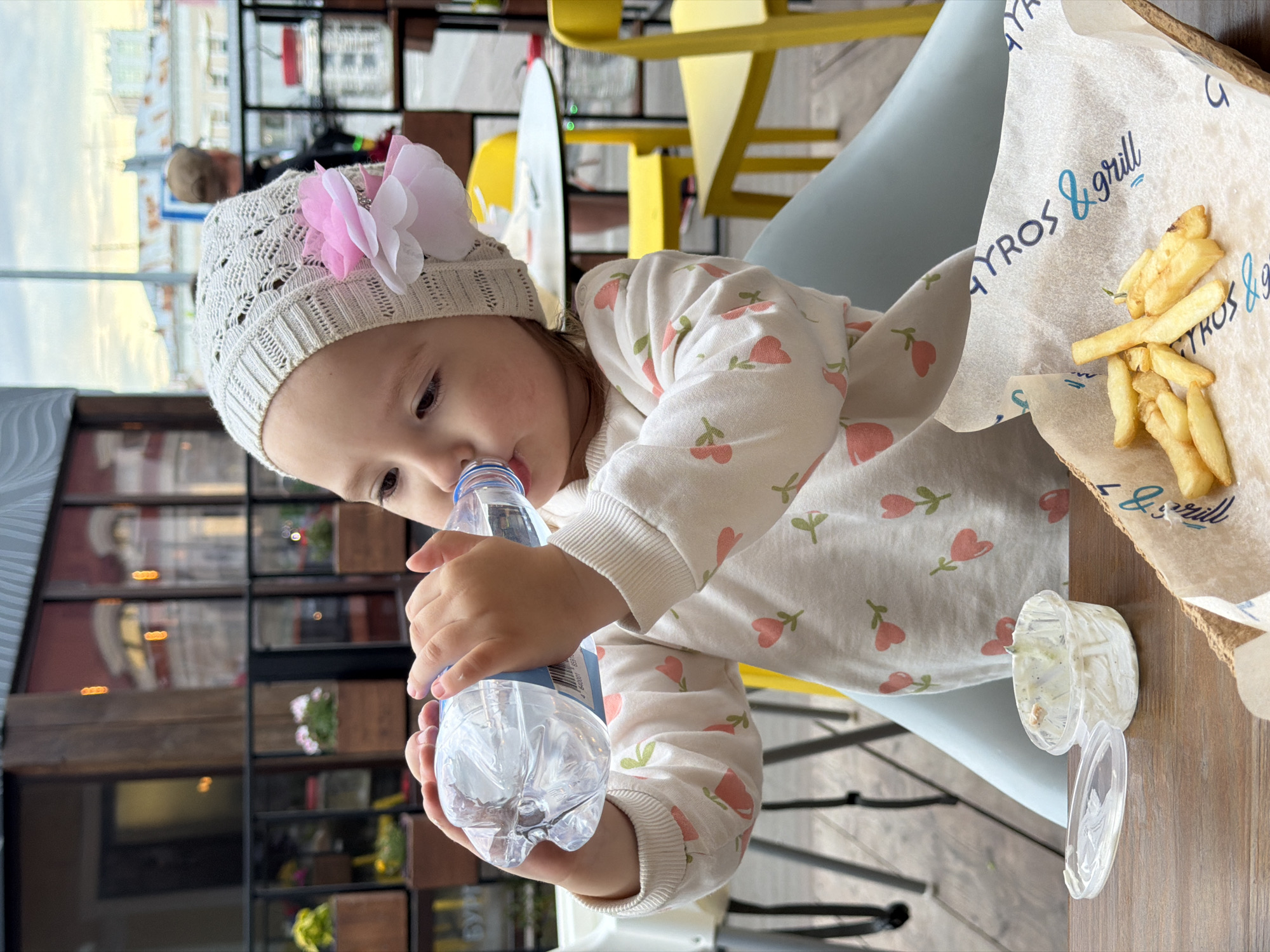 A young girl with a pink and white floral hat is drinking from a clear plastic bottle of water. She is wearing a light-colored long-sleeved shirt with a pattern of small pink tulips. The girl is seated at a wooden table, where a small container with white sauce and a basket lined with paper containing french fries are visible. The background shows an outdoor seating area with yellow chairs and tables. In the distance, buildings and a person walking can be seen. The setting appears to be a cafe or restaurant.