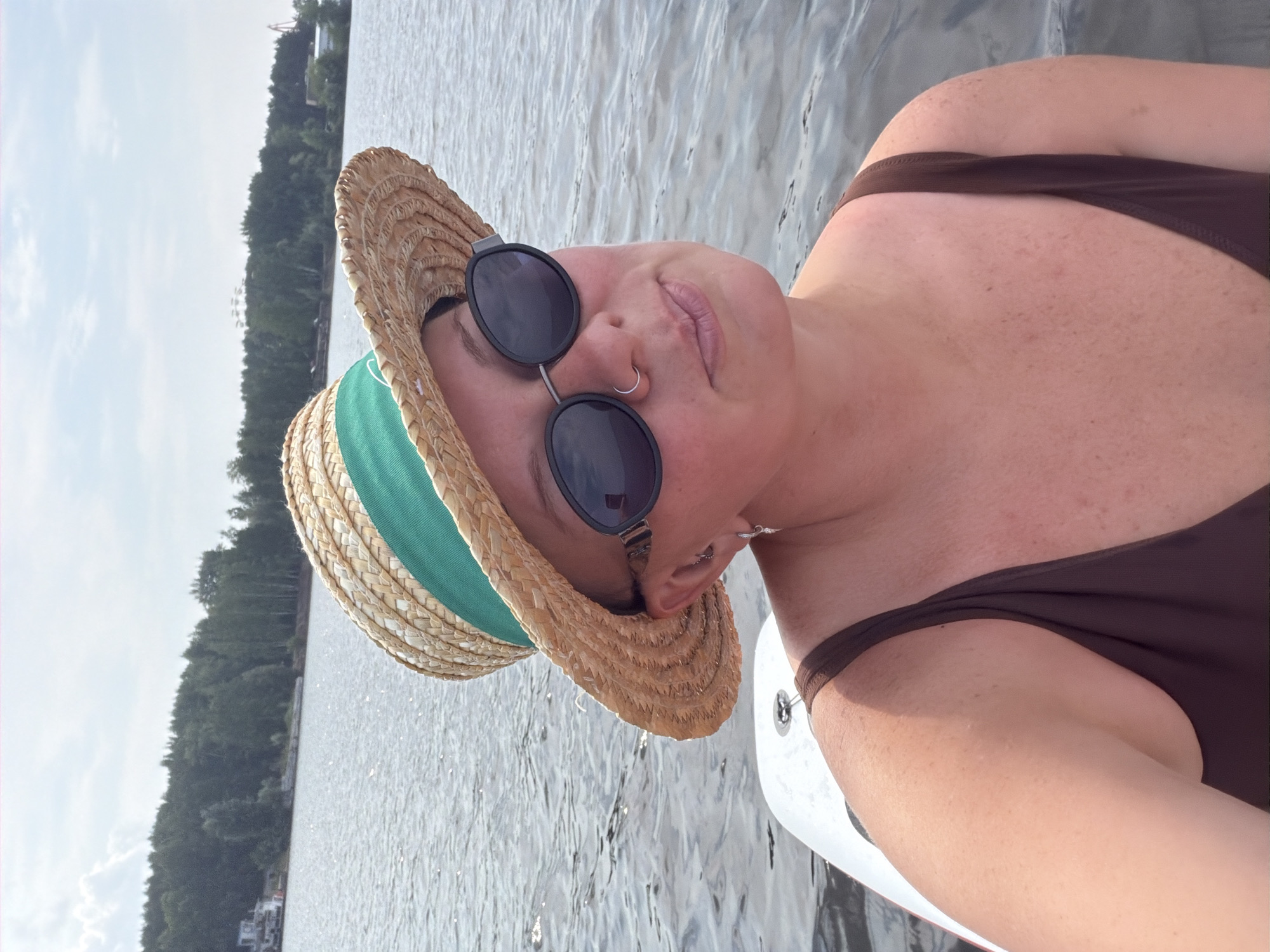 A woman with fair skin, freckles, and brown hair is positioned in the foreground of the image, looking slightly to her right and upwards. She is wearing a brown swimsuit, a straw hat with a green ribbon, and dark sunglasses. A silver hoop earring is visible in her left ear, and a silver nose ring is in her septum. She is on what appears to be a white paddleboard in a body of water, with ripples suggesting a slight chop.
The background shows a calm body of water reflecting the sky. On the far side of the water is a tree-lined shore with some buildings and structures visible. In the distance, a Ferris wheel-like structure and what might be industrial equipment are discernible on the horizon. The sky is partly cloudy with a bright, overcast sun.
The location is identified as the Kama River in Chaikovsky, Russia. The water is likely the Chaikovsky Reservoir, a section of the Kama River that flows through the city and is known for recreational activities. The presence of the paddleboard and the woman's attire suggest a leisure activity on the water. The landscape, with its dense coniferous trees and the distant industrial elements, is characteristic of the region.