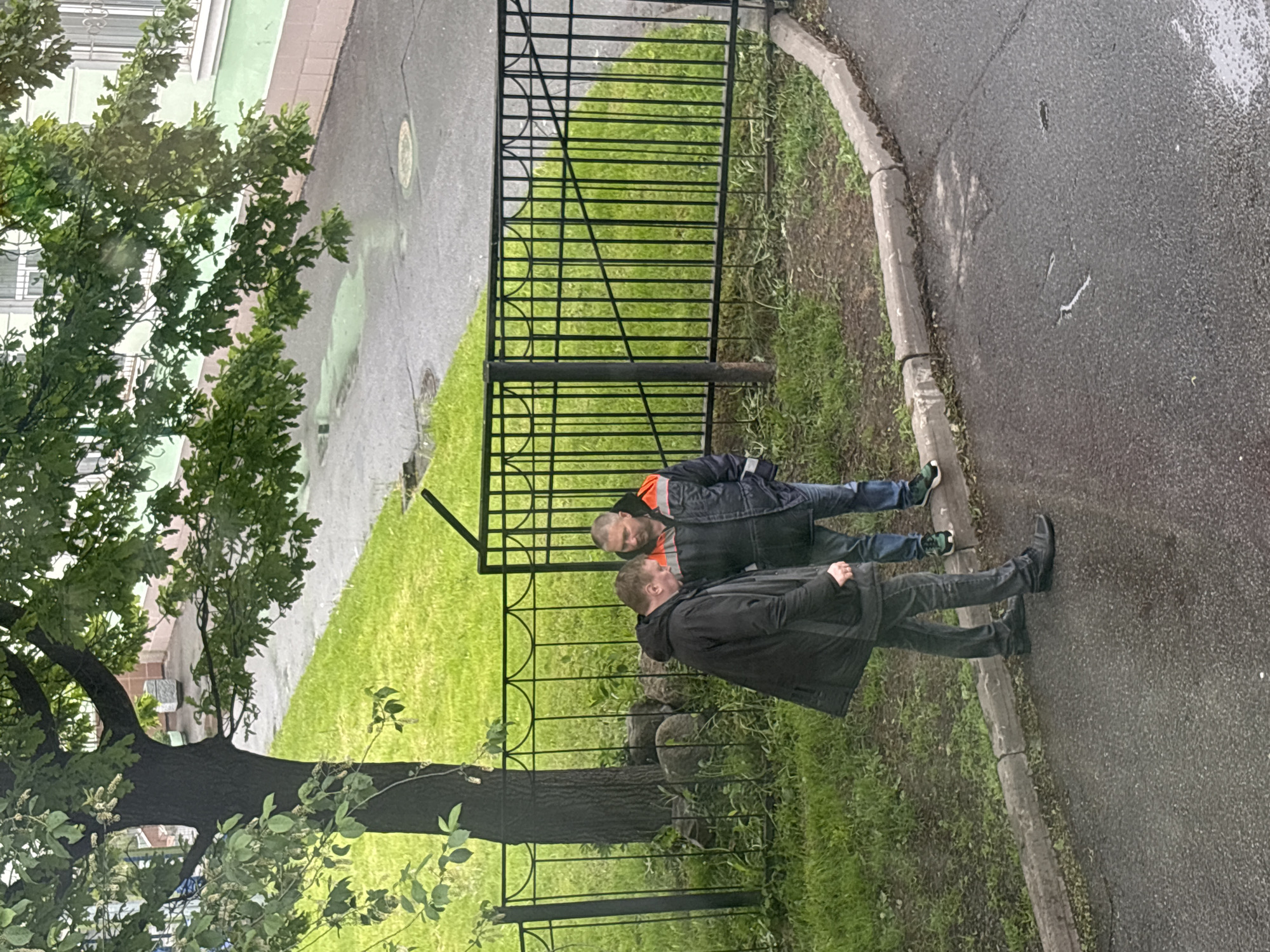 Two adult men stand on a paved sidewalk next to a grassy area enclosed by a black metal fence. One man wears a dark jacket and jeans, while the other wears a black jacket with orange accents. A tree is located on the left. The image appears to be taken from a high vantage point. The location is Saint Petersburg, Russia.