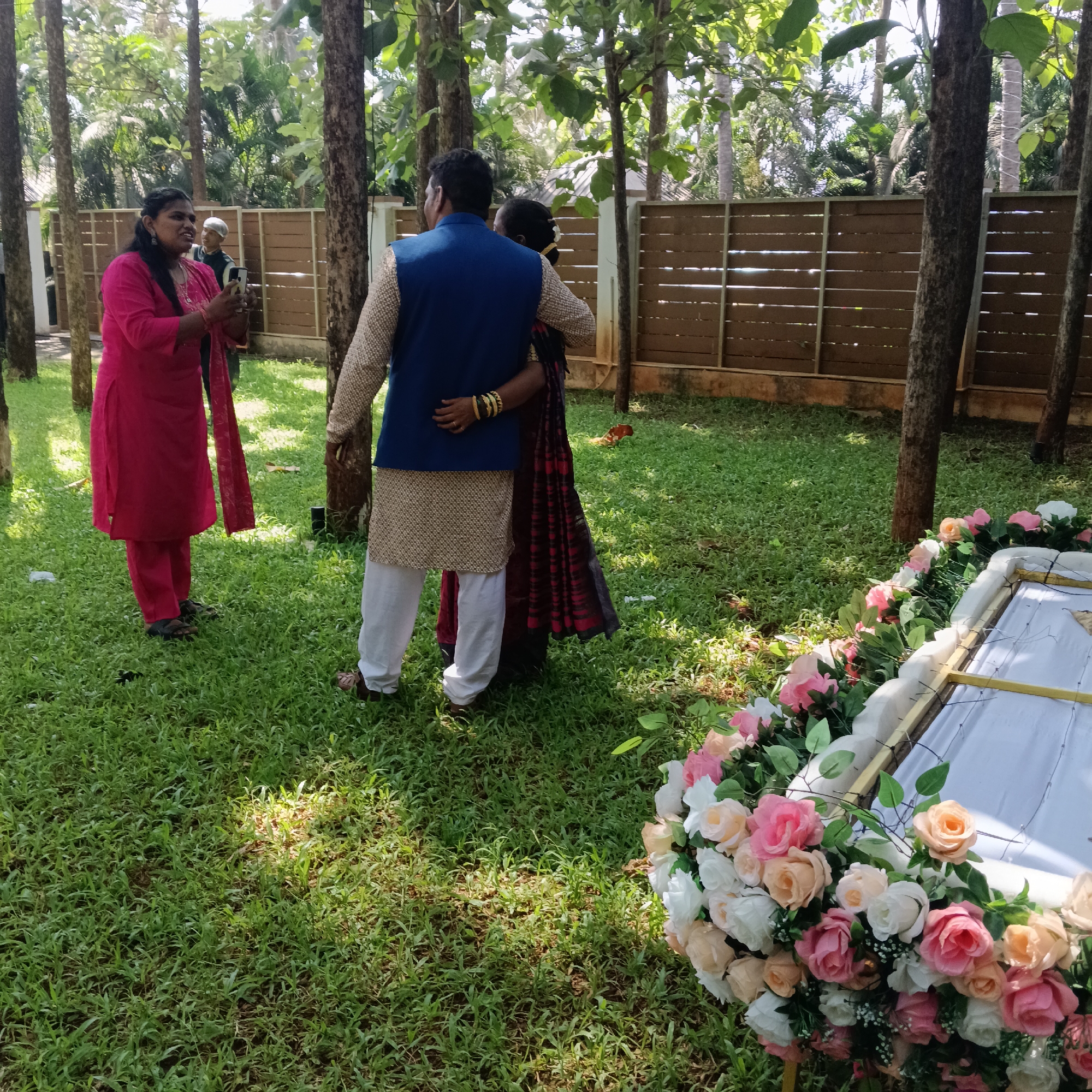 The image depicts a scene in a grassy yard with several tall, thin trees scattered throughout. A wooden fence is visible in the background.
Three individuals are present. On the left, a woman in a bright pink salwar kameez holds a smartphone, seemingly taking a photograph. To her right, a man with his arm around a woman. He wears a beige kurta, white pants, and a navy blue vest. The woman wears a saree with red and black patterns.
In the foreground, a white casket is partially visible, adorned with a large arrangement of pink and white roses.
The scene is likely in Mapusa, India, based on the location details. The nature of the scene suggests it might be related to a memorial or funeral.