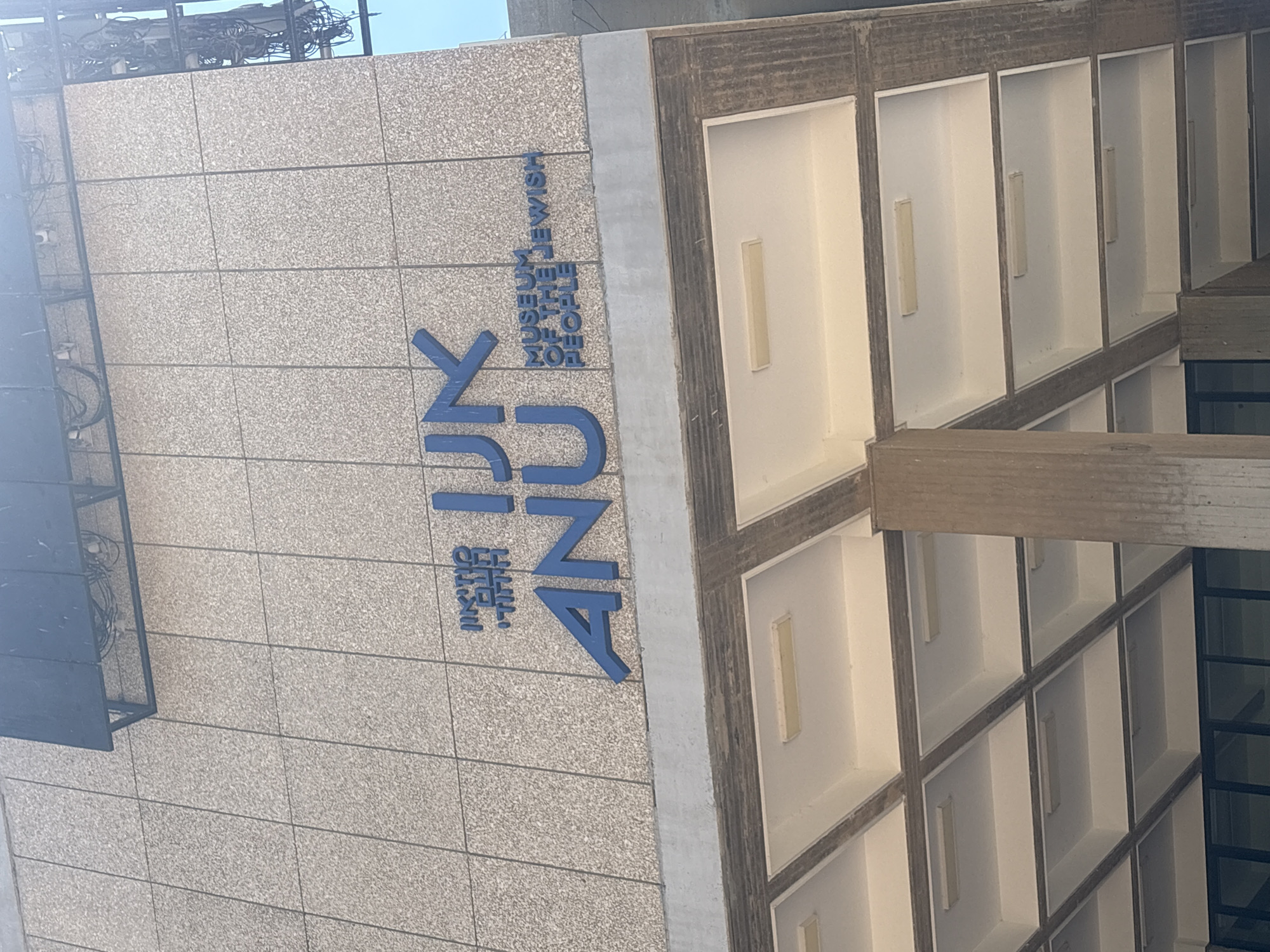 The image shows the exterior of the ANU Museum of the Jewish People in Tel Aviv, Israel. The museum's name is prominently displayed on the building's facade in large blue letters. Above the blue letters, smaller Hebrew text is also visible. The building's exterior is clad in light-colored stone tiles. Below the sign, a covered walkway with recessed lighting is visible. The overall impression is of a modern institutional building.
