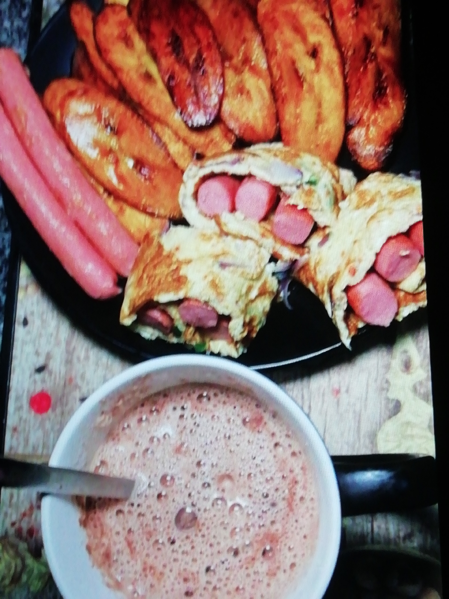 A plate of food is centered in the frame, consisting of fried plantains, two sausages, and pieces of omelet filled with sausage. Below the plate, a cup of a brown, frothy liquid, possibly chocolate milk, is visible with a spoon inside. The setting is a wooden table, and the image is taken indoors. There is no information provided to determine the location is in Enugu, Nigeria. No people are in the frame. The focus is on the food and drink.