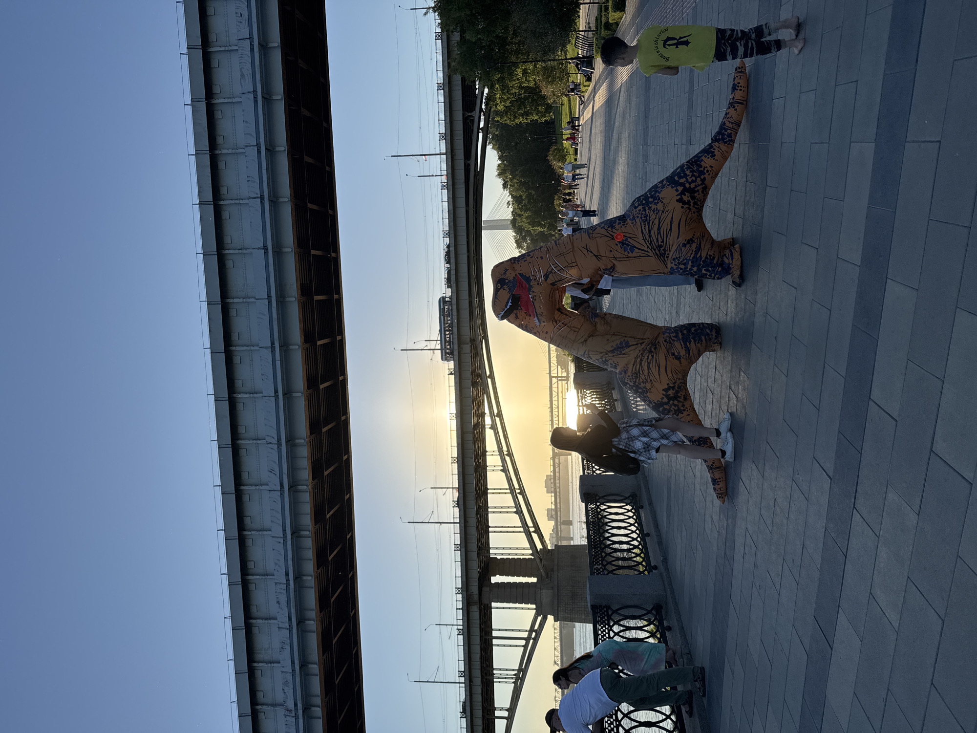 In Novosibirsk, Russia, people in dinosaur costumes walk along a paved promenade by a river. The scene appears to be an outdoor event or gathering. The sun is setting, casting a warm glow across the sky and reflecting on the water. A large bridge, possibly a railway bridge, dominates the background, with another more distant bridge visible further down the river. Trees and some park benches are also present on the promenade. The overall atmosphere seems relaxed and fun, with families or friends enjoying the evening.
