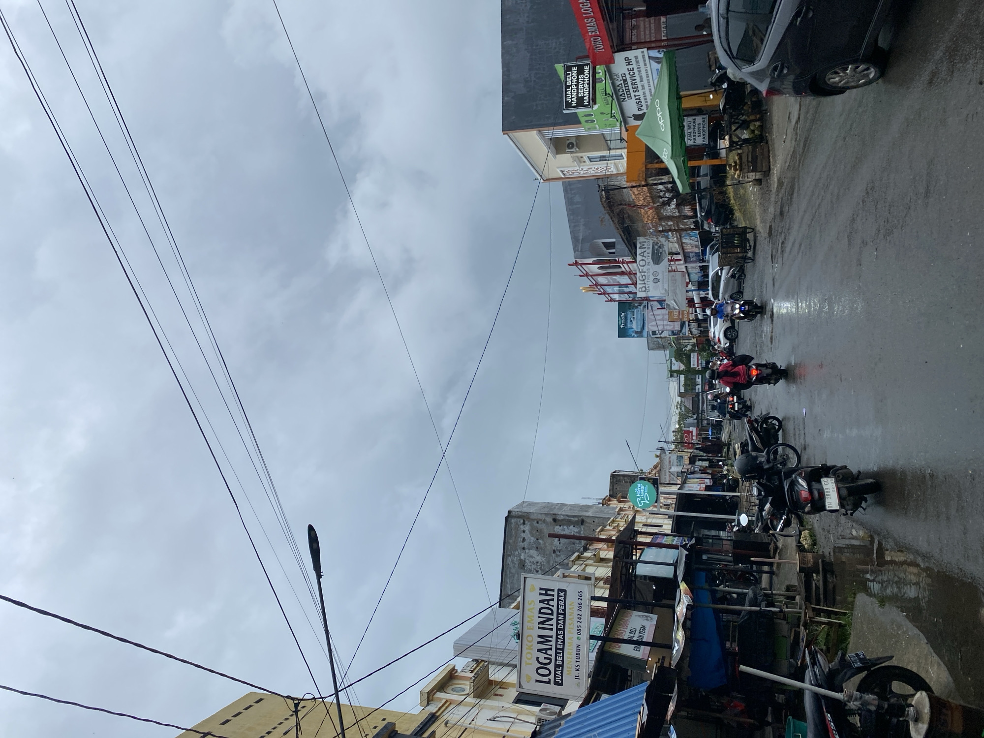 The image depicts a street scene in Ta', Indonesia, under an overcast sky. The street is wet, indicating recent rain, and several motorcycles are visible, some being ridden and others parked along the roadside. Buildings line both sides of the street, featuring various shops and businesses. Signage indicates businesses such as "TOKO EMAS LOGAM INDAH" (Gold Shop Metal Beautiful), which appears to be a jewelry store selling gold and silver, and "NADA Cell" which offers cellphone sales and service. There are also advertisements for mattresses and other products. A few pedestrians are present, and several cars are parked or driving on the street. The overall atmosphere is that of a bustling urban commercial area during a cloudy day.