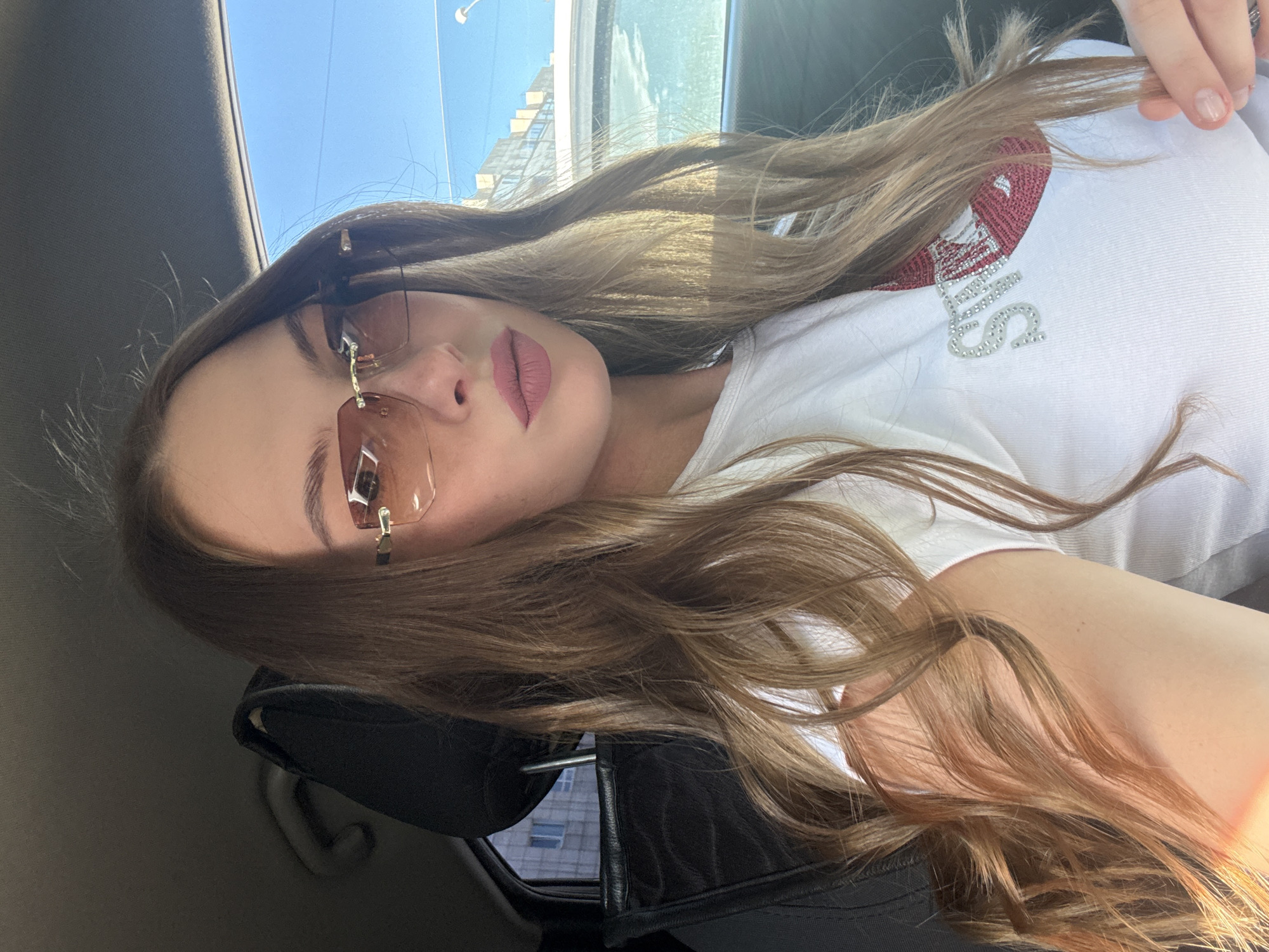 The image shows a young Caucasian woman with long, wavy, light brown hair, wearing rimless sunglasses with tinted lenses and a white t-shirt adorned with sparkly embellishments. She is seated in the driver's seat of a car, with the car's interior visible, including a headrest and part of the dashboard. The background, seen through the car window, suggests an urban environment with buildings and a bright blue sky, consistent with a location in the city of Perm, Russia. The lighting indicates it is daytime, likely sunny.