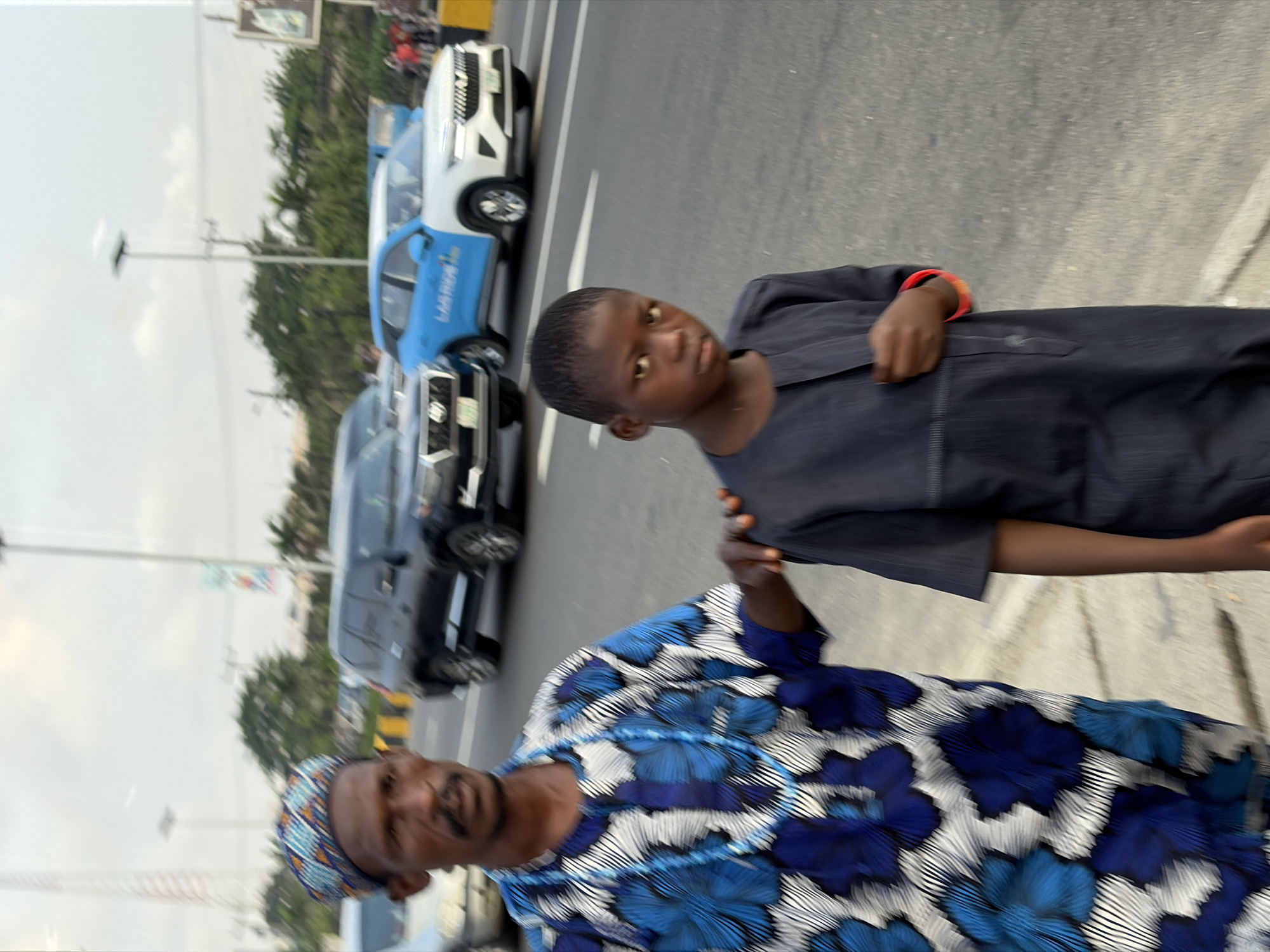 The image shows two individuals, an adult male and a young male, standing on a paved road in Igboefon, Nigeria. The adult male, on the left, wears a patterned blue and white traditional garment and a patterned headwrap. He has a mustache and is looking towards the camera. The young male, on the right, is wearing a dark, solid-colored garment and is also looking towards the camera. His left hand is resting on his chest. The adult male has his right hand on the young male's shoulder. In the background, there is a street with several vehicles, including a black SUV and a white and blue car with "LAGBUS" written on it. Trees and streetlights are also visible. The sky is overcast.
