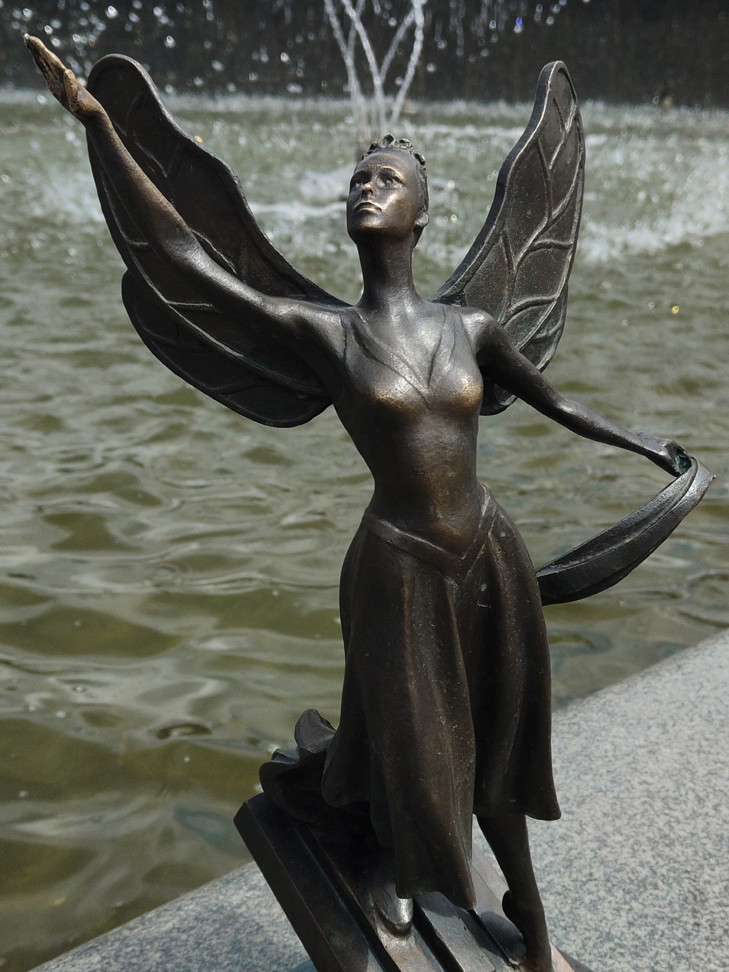 A bronze statue of a winged female figure stands prominently in the center of the frame, positioned at the edge of a body of water. The figure appears to be a fairy, angel, or mythical being, rendered in a dark, weathered bronze with a subtle sheen on its raised surfaces.
The statue is captured in a dynamic, graceful pose, with its left arm reaching upwards and outwards as if in flight or greeting, its hand open. Its right arm is extended to the side, holding a flowing piece of drapery or ribbon that trails behind it. The figure's head is tilted slightly back, and its gaze is directed upwards, conveying a sense of aspiration or wonder. It features a delicate facial structure and appears to wear a small crown or intricate headpiece.
Large, finely detailed wings, resembling stylized leaves or insect wings with visible veins, extend from its back, contributing to the ethereal appearance. The figure is clad in a flowing gown or dress, with soft folds and an elegant V-neckline. Its posture suggests movement, with one leg slightly bent.
In the background, a body of water, likely a fountain or pond, stretches out, its surface rippled and a greenish-brown hue. Several white, frothy jets of water are visible, some blurred into bokeh, indicating an active fountain. The statue is mounted on a gray, speckled stone or concrete base, which forms a clear boundary with the water. The scene is brightly lit, suggesting it is daytime under clear weather conditions, casting subtle shadows and highlighting the bronze.
There is no visible text in the image.