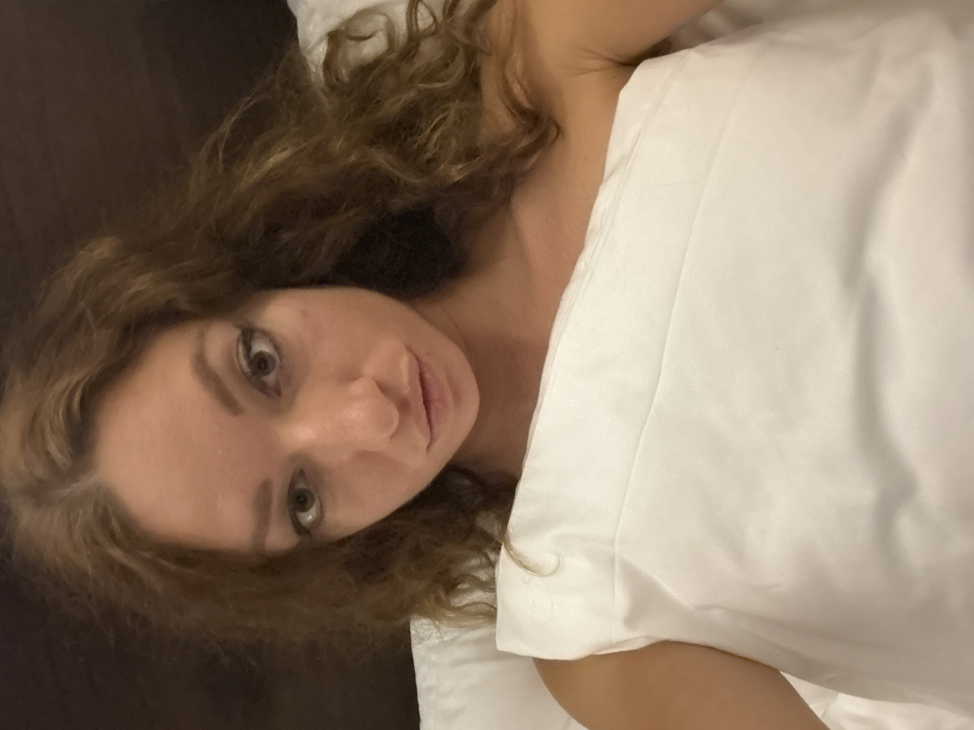 A woman with long, wavy brown hair lies in bed, her head resting on a white pillow. She is looking directly at the camera with her lips slightly pursed. Her left shoulder is bare, and she is covered by white bedding. The background is dark and out of focus, suggesting an indoor setting. There are no visible landmarks to indicate a specific location in Nam Hoa Lư Ward, Vietnam.