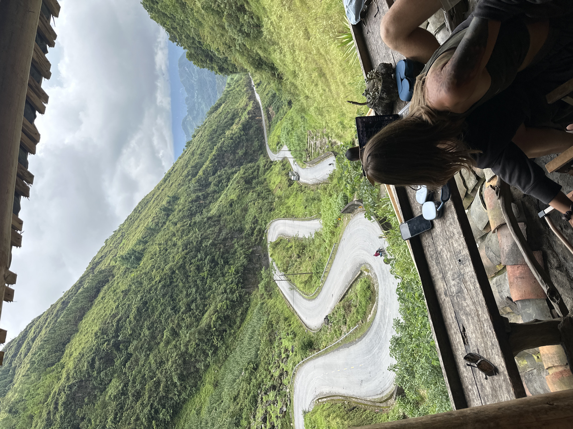 A scenic overlook captures a meandering road carved into a lush, green mountainside in the Yên Minh Commune, Vietnam. The road descends into a valley. A portion of the image shows the top of a partially obscured cliff covered in dense vegetation.
Two individuals are seated on a wooden deck, with one facing the view and using a laptop. Various personal items, including sunglasses and shoes, are scattered on the deck. The deck appears to be part of a rustic, open-air structure. The weather appears overcast, with cloudy skies in the distance.