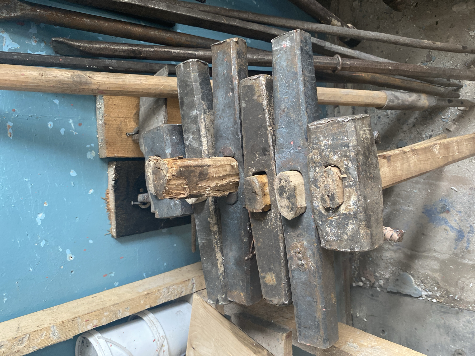 The image depicts a close-up, high-angle shot of a pile of old, heavy-duty tools leaning against a blue, peeling painted wall. The tools are arranged on a wooden surface, with some appearing to be sledgehammers or similar impact tools with thick, worn metal heads and wooden handles. Small pieces of wood are interspersed among the tools. To the left of the tools, a portion of a white bucket and a raw wooden plank are visible. The background wall is textured and shows signs of wear and tear, with chipped paint and some scattered debris on the floor in the foreground. The scene is likely an interior space such as a workshop or storage area. There are no people or animals visible in the image. This image is not specific to Oktyabrsk, Russia.