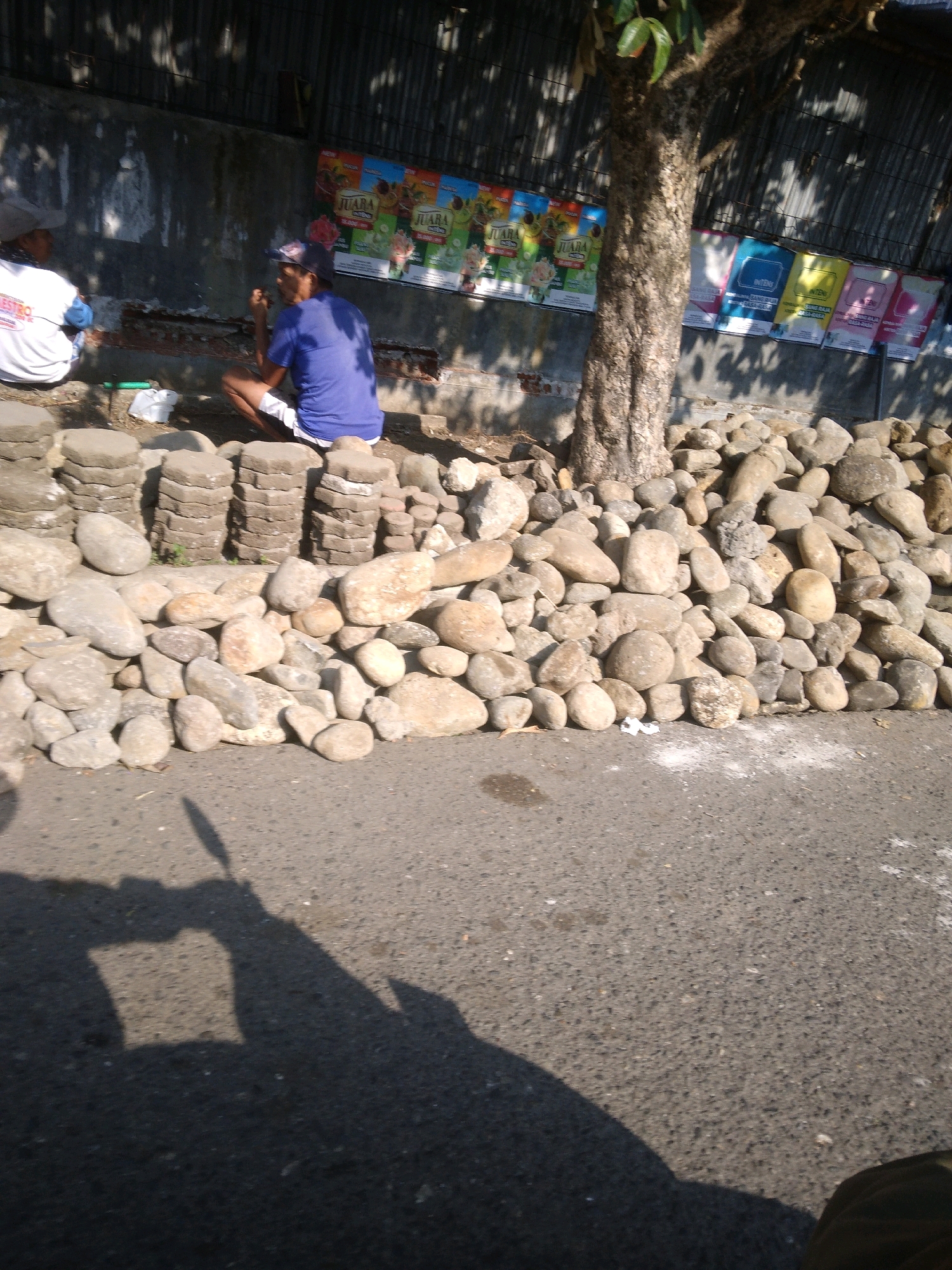 The image shows a street scene in Purwokerto Kulon, Indonesia, with a pile of smooth, grey, rounded stones in the foreground. Behind the stones, a man wearing a blue t-shirt and white shorts is sitting on the ground, holding something to his mouth. To his left, another person wearing a white shirt with red lettering is also sitting. A tree trunk with rough bark is visible behind them, casting shadows on the ground. In the background, a corrugated metal wall is adorned with several colorful posters featuring text and images. The ground is paved with asphalt.