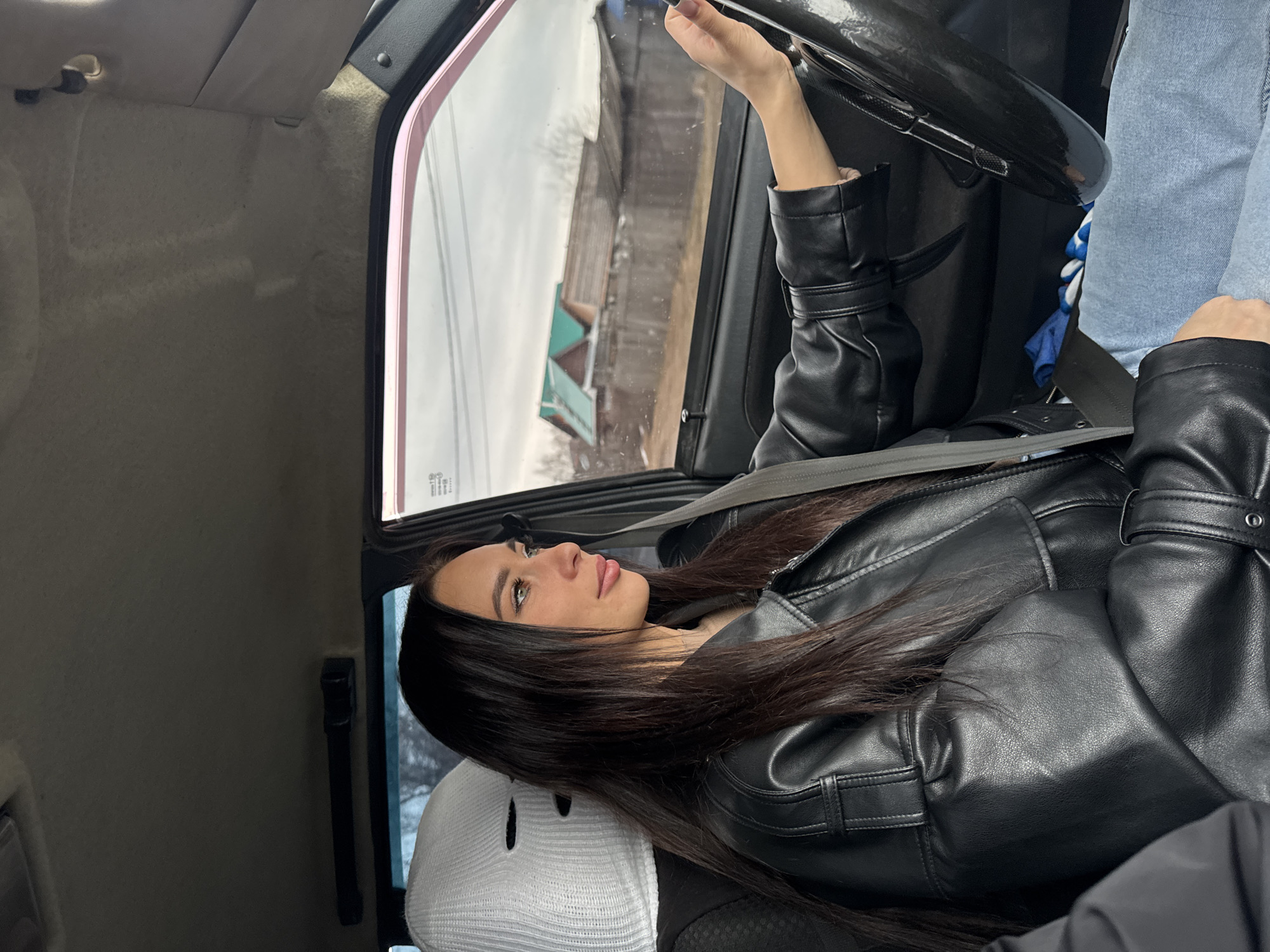 A woman wearing a black leather jacket is sitting in a car, restrained by a seatbelt. She rests her head on a white ski mask placed on the headrest. She looks out the window, where a building can be seen. The photo was taken in Chaikovsky, Russia.