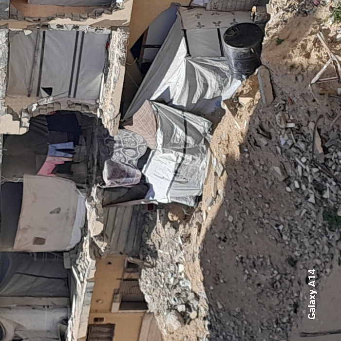 The image displays a scene in Khan Yunis, Palestinian Territory, showing an area with significant structural damage and improvised living structures under clear daylight conditions. In the background, two multi-story buildings exhibit severe damage, with large portions of their facades missing, exposing interior spaces and fragmented structural components. Openings and cavities within these damaged buildings are covered with various textiles and tarpaulins, including white, light grey, and patterned fabrics. Clothes, specifically a pink top and blue trousers, are visibly hanging to dry from an upper-level opening on the left building.
The mid-ground and foreground feature mounds of dry earth, sand, and extensive rubble, consisting of shattered concrete, stones, and other debris. Situated among this detritus and directly beneath the damaged buildings are several temporary shelters. A prominent tent-like structure, constructed from white and grey tarpaulins with horizontal black banding, extends towards the right of the frame. Above and to the left of this main tent, a smaller section of patterned fabric is visible, possibly part of another shelter or awning. A black cylindrical barrel lies horizontally on the ground to the right of the primary tarp structure. No individuals are visible within the frame.