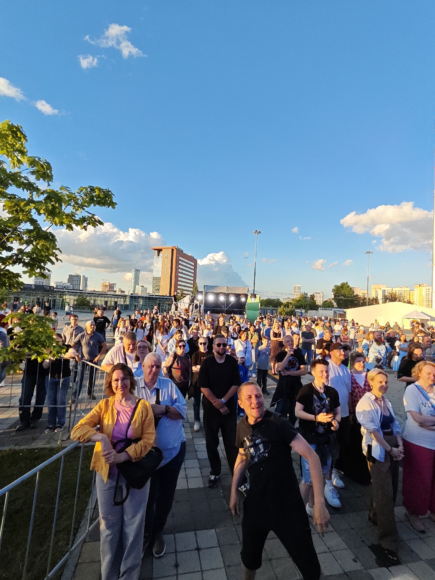 This media file depicts an outdoor event, likely a concert or festival, taking place in Yekaterinburg, Russia. A large crowd of people is gathered, facing a stage with bright lights visible in the background. The sky is a clear blue with scattered white clouds, suggesting fair weather. The time of day appears to be late afternoon or early evening, indicated by the warm, golden light casting long shadows.
The crowd consists of individuals of various ages and backgrounds, dressed casually. Many people are looking towards the stage, some with excited expressions. In the foreground, a woman in a yellow jacket and gray pants is leaning on a metal barrier, smiling at the camera. Behind her, a man in a blue shirt is also visible. Further back, a man in a black t-shirt and black pants is captured mid-movement, seemingly dancing or interacting with the crowd.
The setting includes a paved area where the audience stands, and a metal fence separating them from the immediate area in front of the stage. In the distance, urban structures, including buildings of different heights and a tall tower, are visible against the skyline. The overall atmosphere appears to be lively and engaging. There is no visible text in the image.