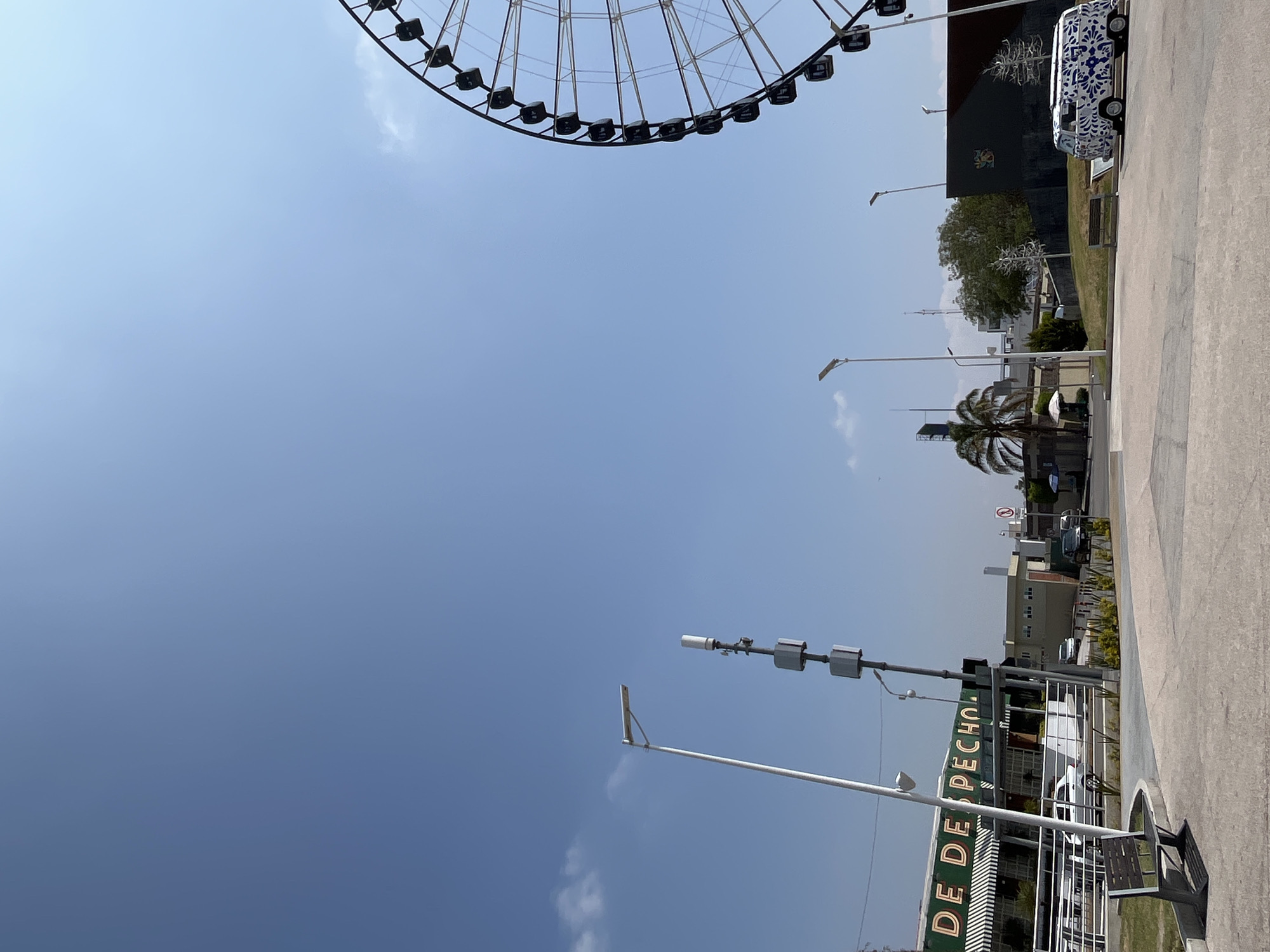 The image shows a Ferris wheel partially visible at the top, against a bright blue sky with some scattered clouds. Below, a street scene is present, featuring a tall sign that says "DE DE DESPECHO". Several lampposts are visible, along with buildings, trees, and vehicles. A parking area is also present on the right side of the image. The location is the Parque del Teleférico in the city of Puebla, Mexico.