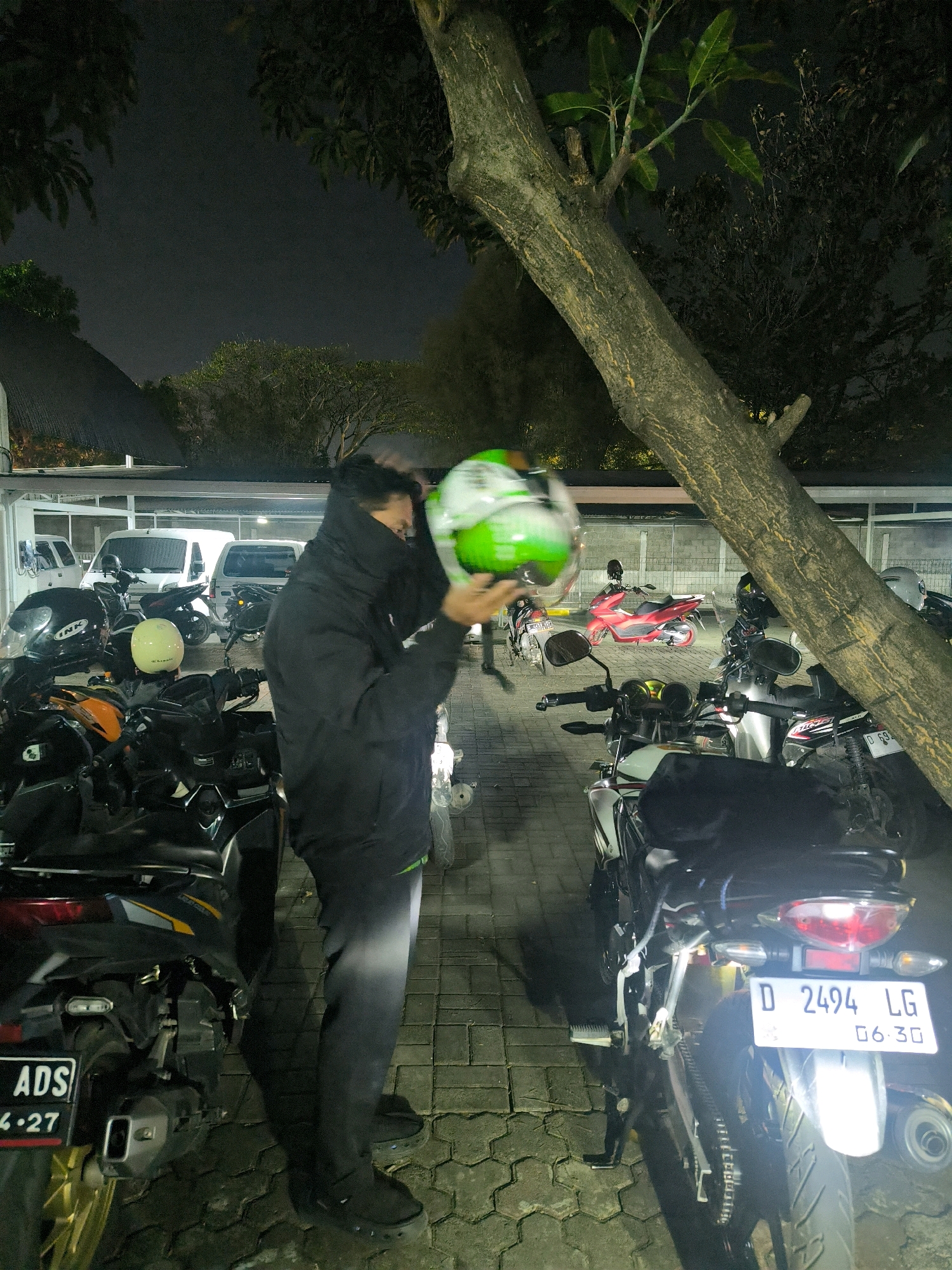 A person wearing a black jacket and dark pants is holding a green and white helmet while standing next to parked motorcycles at night. The scene appears to be an outdoor parking area, possibly a motorcycle parking lot or a street, with paved ground made of hexagonal stones. Several motorcycles are visible, including a red scooter and various other models parked closely together. In the background, white vans are parked under a covered structure. A large tree trunk with textured bark diagonally crosses the frame, obscuring parts of the scene. The lighting suggests it is nighttime, with artificial lights illuminating the area. License plates are visible on some of the motorcycles, one reading "D 2494 LG" and another partially visible plate reading "ADS" followed by numbers. The environment is consistent with a location in Bandung City, Indonesia, characterized by the presence of motorcycles as a common mode of transportation and the type of vehicle and license plate observed.