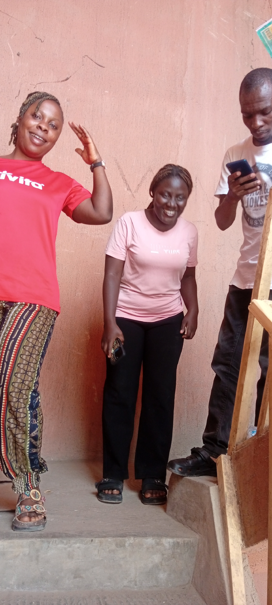 Three individuals, two women and one man, are pictured indoors against a textured, light brown or peach-colored wall with some visible marks. They appear to be standing on concrete steps or a raised platform, with a wooden structure (possibly part of a railing or frame) on the right side of the image.
On the left, a woman with braided hair smiles widely and raises her right hand. She is wearing a red short-sleeved t-shirt with the text "vivita" visible on it, and distinctive patterned pants in shades of blue, red, brown, and white. Her footwear consists of open-toed sandals with ornate, gold-colored buckles.
In the center, another woman, also with braided hair, is captured mid-laugh, her face radiating joy. She wears a light pink short-sleeved t-shirt with text that appears to be "YUNE" or similar, black pants, and black open-toed sandals. She holds a smartphone in her right hand.
On the right, a man is partially visible, from his chest down. He is wearing a white t-shirt with "JACK & JONES" and "ORIGINAL" printed on it, and dark pants. He is focused on a smartphone held in his hands. His footwear consists of dark, somewhat robust shoes, possibly clogs or work shoes.
The scene conveys a casual and cheerful atmosphere, particularly from the smiling and laughing women. The lighting suggests it is daytime. The location is an everyday setting in Fagge C, Nigeria.
