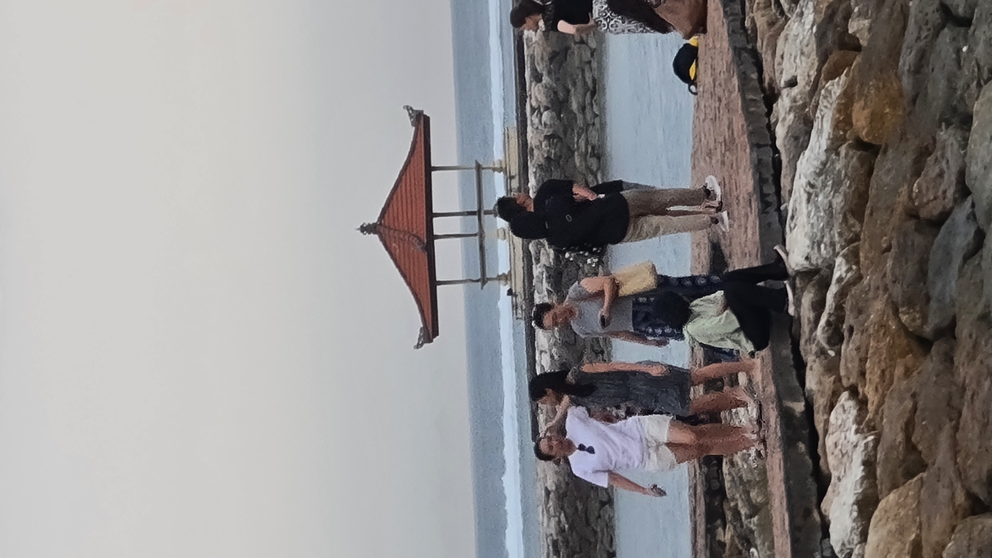 This media file depicts a group of people standing along a stone embankment next to a body of water, likely the ocean. The scene appears to be set in Denpasar, Indonesia, indicated by the presence of a traditional Balinese pavilion with a multi-tiered, red-tiled roof. The sky is overcast, suggesting a cloudy or possibly twilight time of day.
The subjects are varied. Several individuals are visible, dressed in casual attire. One couple is in the foreground, with the male wearing a black jacket and the female in a patterned dress. Further down the embankment, other people are standing, some appearing to be tourists taking in the view or posing for photos. One individual is wearing a white t-shirt and shorts, while another is in a light green top and dark pants. A person in a grey t-shirt and patterned shorts is holding a phone. The overall mood seems relaxed and leisurely, with people enjoying the coastal scenery.
The setting is characterized by a rugged stone wall forming the edge of the land, with a pathway or walkway behind it. The water beyond is calm and appears to be a light blue-grey. The Balinese pavilion stands as a distinct architectural element, contributing to the cultural context of the location. There is no visible text in the media file. The image captures a moment of everyday life in a scenic coastal area, possibly a popular tourist spot.