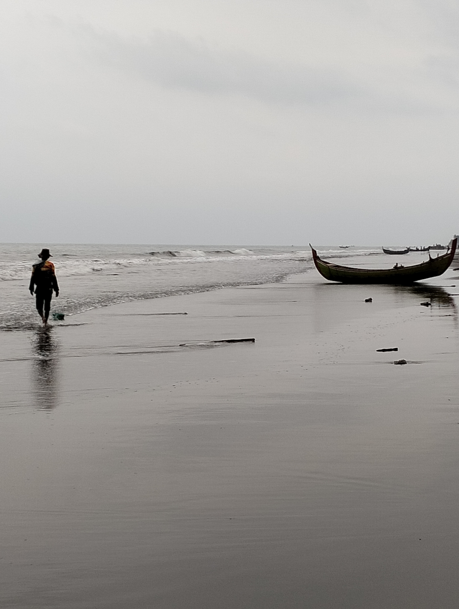In Peuneulet Baroh, Indonesia, a solitary figure walks along a wet, dark sand beach under an overcast sky. The person, silhouetted and wearing a hat, walks towards the water's edge, their reflection shimmering on the wet sand. The ocean stretches to the horizon, with gentle waves lapping the shore. Several fishing boats are pulled up onto the beach, the largest of which is a long, slender vessel with an upturned bow and stern, painted a dark color. Scattered debris, possibly driftwood, lies on the sand. The overall mood of the image is somber and tranquil, with the muted colors and diffused light contributing to a sense of quiet contemplation.