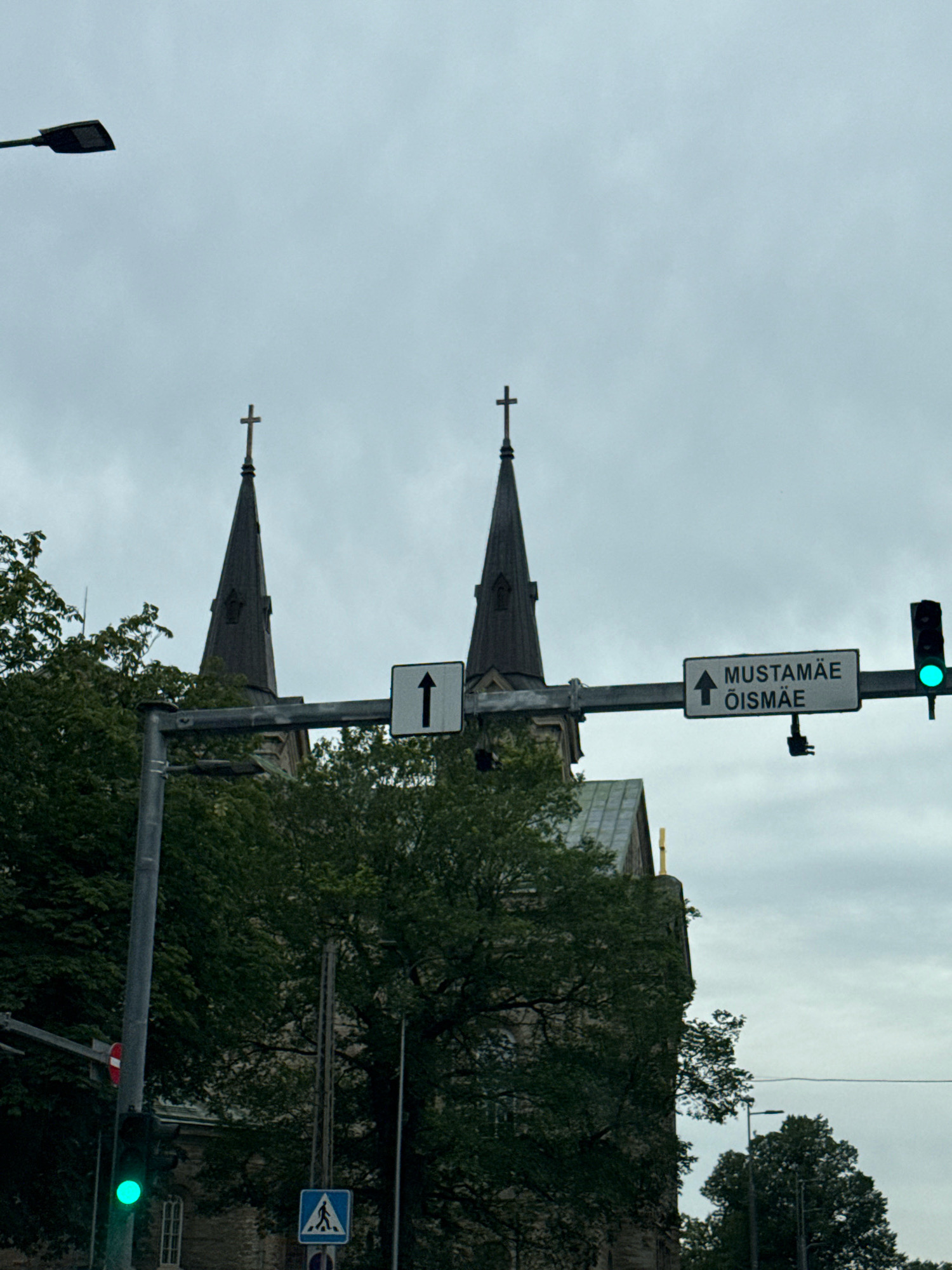 The image depicts an overcast day in Tallinn, Estonia, with a focus on a street intersection. Dominating the background are two spires of a church, likely St. Olaf's Church, given its prominent silhouette and its typical location within the city. A street light is visible in the upper left corner. A traffic light hangs above the road, currently displaying a green light. To the right of the traffic light, a street sign indicates "MUSTAMÄE ÕISMÄE" with an upward arrow, directing traffic towards these districts. In the lower left quadrant, a traffic light displays a green signal, and a pedestrian crossing sign with a blue background and white pictogram is visible below. Lush green trees surround the church and parts of the street, suggesting a relatively green urban environment. The overall scene conveys a typical urban setting in Tallinn.