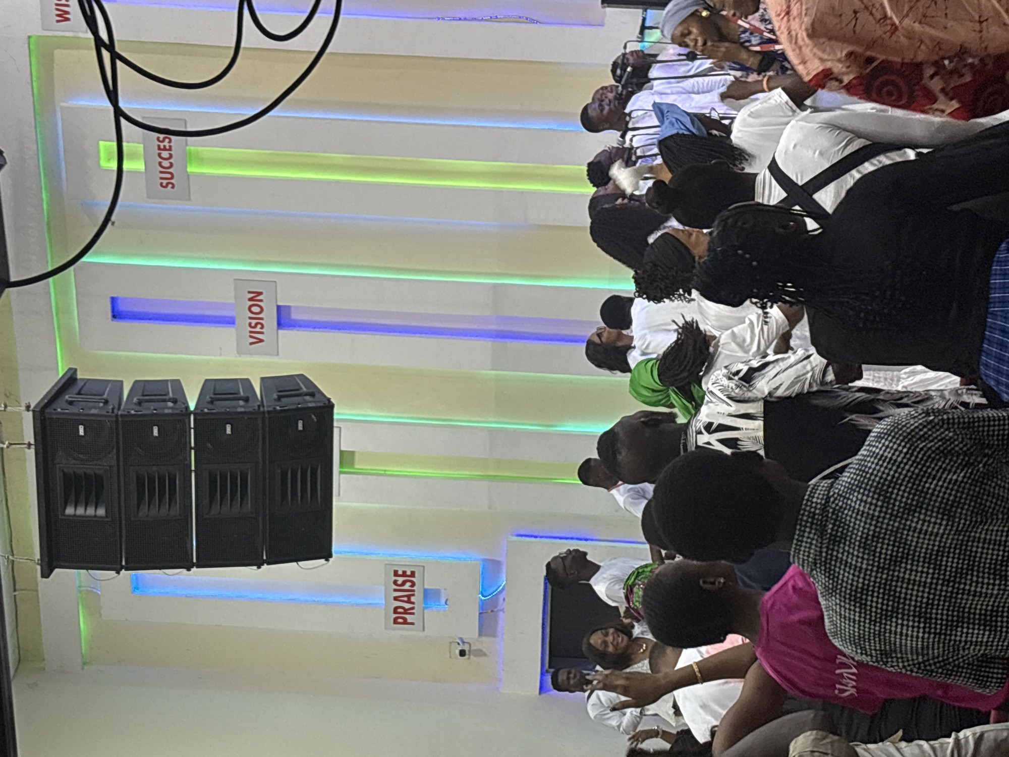 A vertical eye-level shot captures a gathering of people indoors in Lugbe, Nigeria. Most of the attendees wear white clothing, with a few interspersed in other colors such as pink and black. The room is illuminated with alternating blue and green lights along the walls, which also have signs that read "PRAISE," "VISION," "SUCCESS," and "WISDOM." Four large, black loudspeakers are mounted on the wall. Black cables run along the ceiling. The focus is on the gathered people, and their interactions are not visually clear. The environment suggests a formal indoor event.