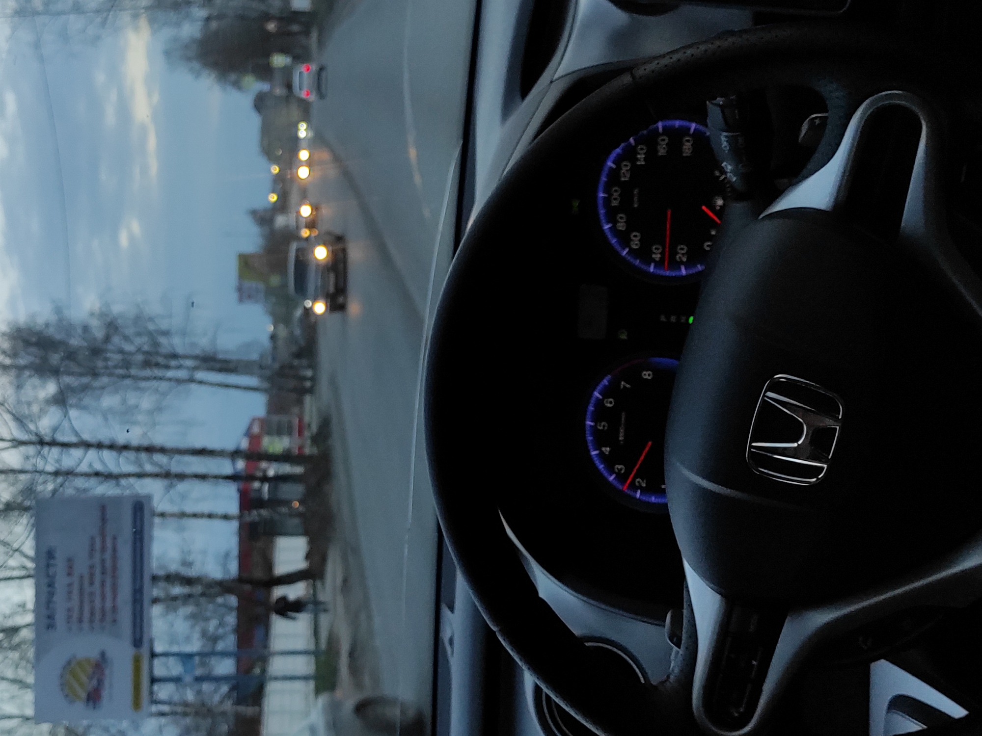 The photo, taken from inside a car, presents a view through the windshield onto a street in Chaikovsky, Russia. The car's steering wheel, prominently featuring the Honda logo, occupies the foreground. The dashboard gauges are illuminated, indicating the car is running. Outside, other vehicles are visible on the road, their headlights on. A sign in Russian advertising spare parts ("ЗАПЧАСТИ") stands on the side of the road. Bare trees line the street. The sky is overcast.