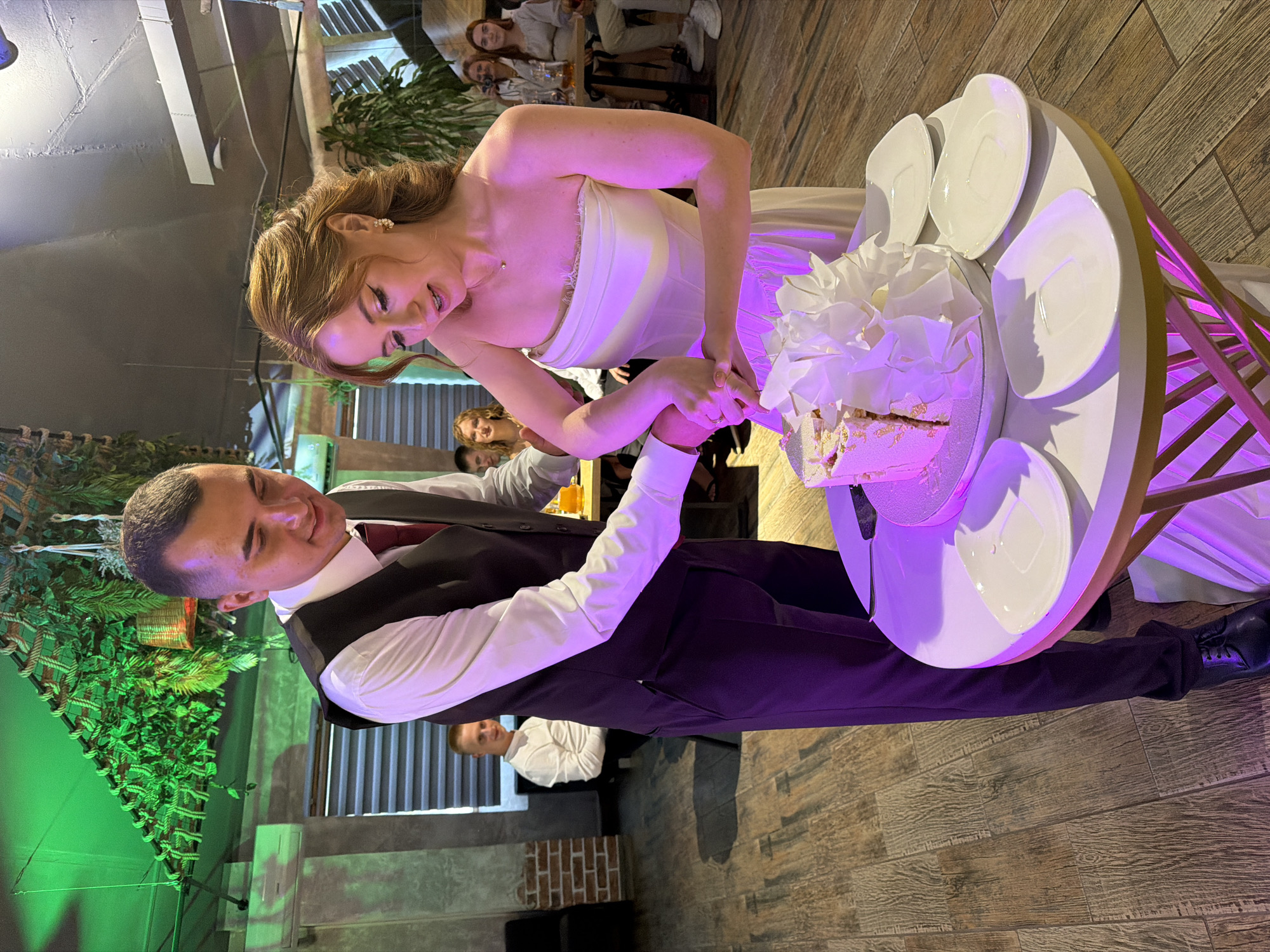 At a wedding reception in Chaikovsky, Russia, a bride and groom are cutting a multi-tiered cake. The bride, wearing a strapless white gown and gold earrings, holds a knife with her groom, who is in a white dress shirt, vest, and burgundy tie. The cake, decorated with white frosting and a large white flower, is on a white circular table with gold trim. Several white plates and a cake server are also on the table. The room features wooden floors, exposed brick, and lush green decorations hanging from the ceiling, illuminated by green and purple lights. Guests are visible in the background, seated at tables.