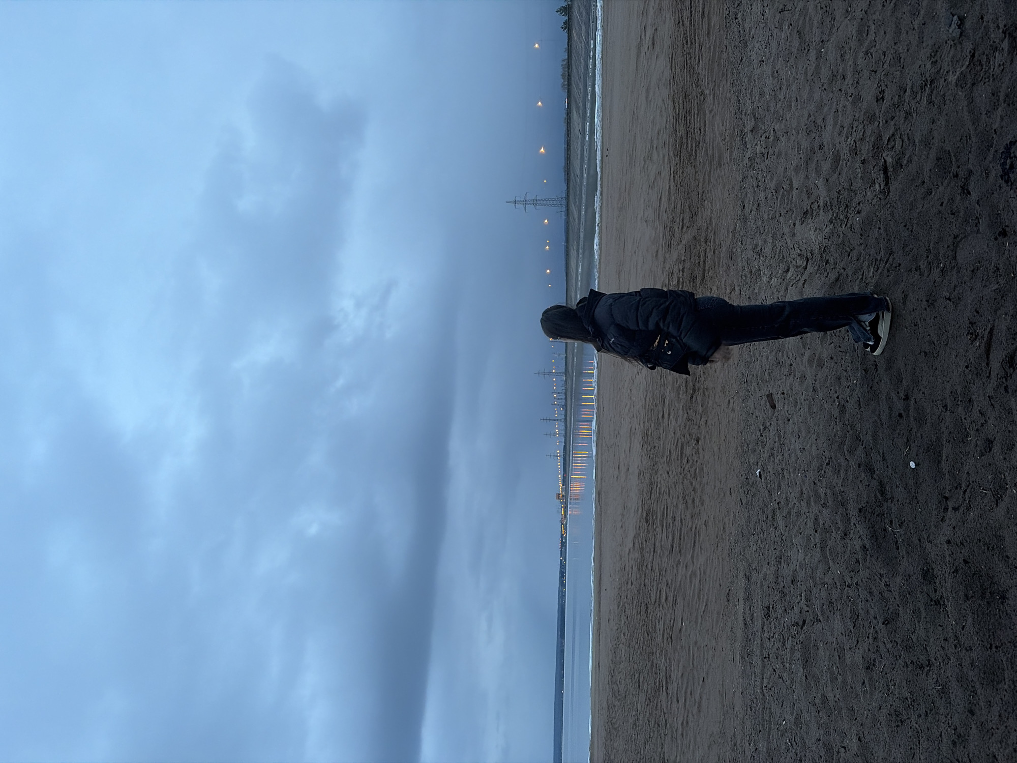 The image depicts a solitary figure standing on a dark, sandy beach at dusk or night in Noviy, Russia. The overall scene is moody and expansive, characterized by a vast, overcast sky filled with dark blue and grey clouds.
A person, viewed from behind, stands on the right side of the frame, facing towards the water and the distant horizon. They are dressed in warm attire, including a dark puffer jacket or coat, dark trousers, and dark sneakers with white soles. Their dark hair is visible beneath the hood or collar of their jacket.
To the left of the beach is a large body of water, reflecting the dim light from the sky and the artificial lights in the distance. Along the far shoreline, a series of bright, yellow-orange lights are visible, creating elongated reflections on the water's surface. These lights suggest an urban or industrial area in the distance. Several tall structures, appearing to be power line towers, stand silhouetted against the twilight sky along the distant coast.
The time of day is clearly evening or night, indicated by the darkness of the sky and the prominent artificial lights. The weather appears overcast and potentially chilly, given the person's warm clothing. The scene evokes a sense of quiet contemplation or solitude. No visible text is present in the image.