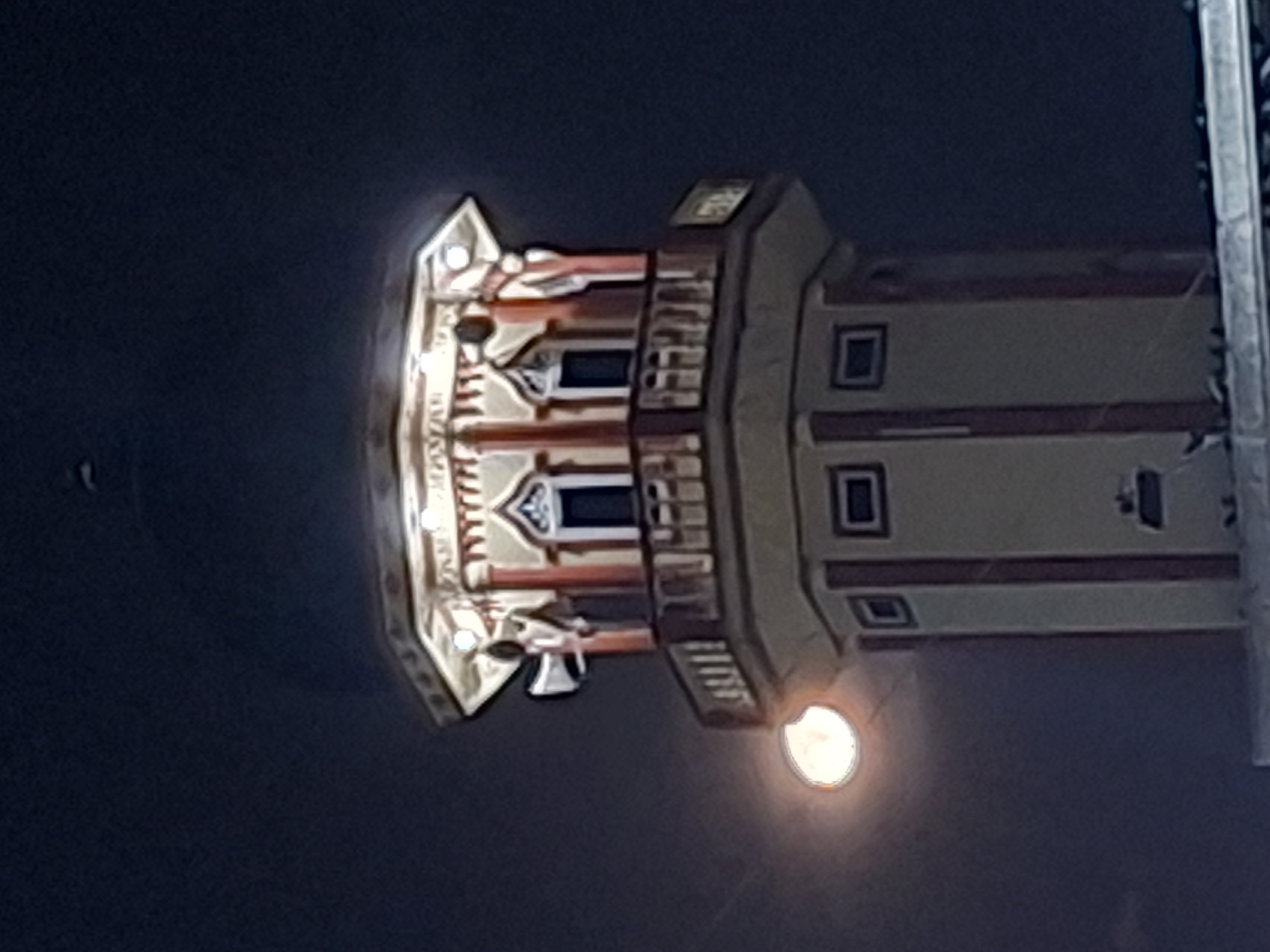 The image shows a tall, brightly lit structure at night. The upper portion of the structure has ornate details and windows with arch-shaped ornamentation. Below the ornate section is a simpler, multi-story section with square windows and a light source illuminating its base. The structure appears to be located in Keudeteupinpunti, Indonesia.