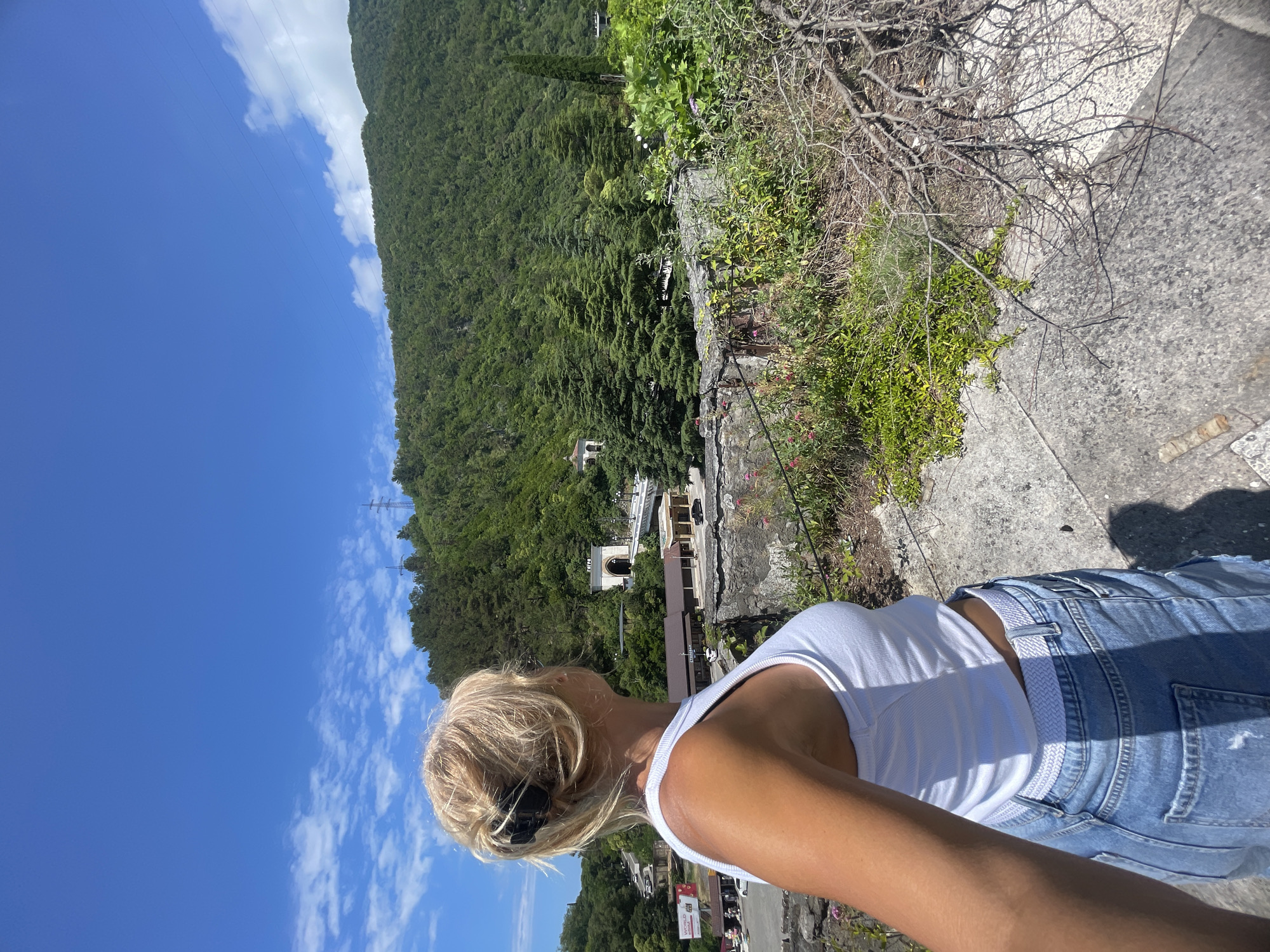 In the city of Abaata, Abkhazia, a blonde woman with her hair pulled back stands on a stone ledge, looking out at a lush, green mountainous landscape. She is wearing a white tank top and distressed denim shorts. In the background, a town or village is visible nestled at the foot of the mountains, with buildings, roads, and vehicles. A prominent feature is a tunnel entrance, possibly for a road or railway, carved into the mountainside, adorned with architectural detailing. The sky is a vibrant blue with scattered white clouds. The foreground is dominated by rough stone, dry branches, and low-lying green vegetation.