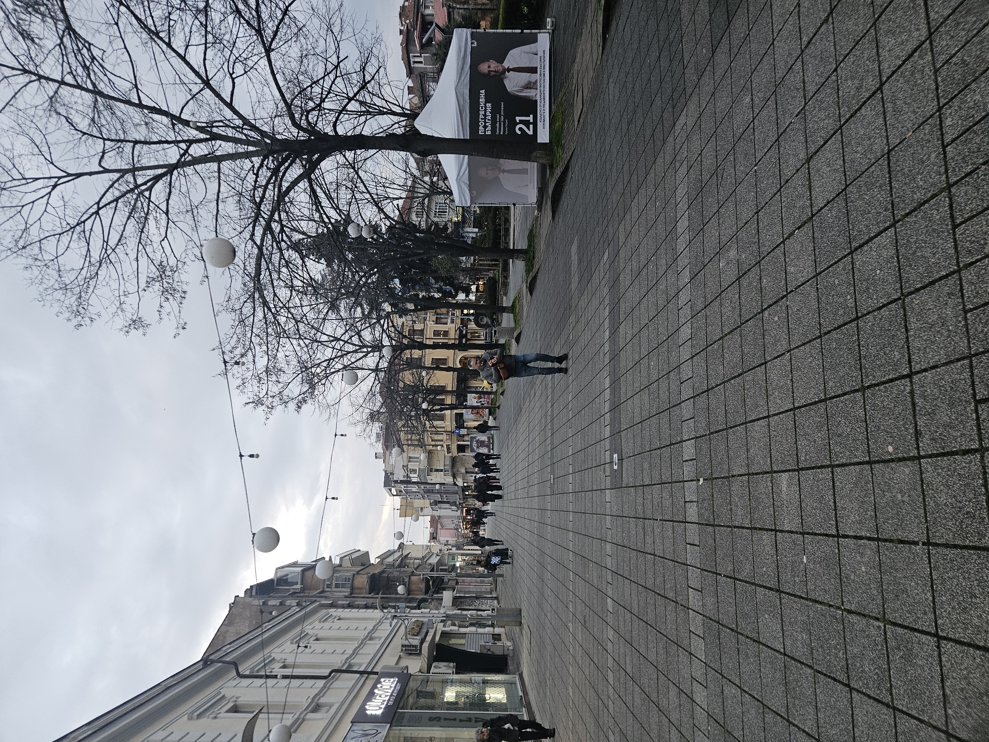 The image captures a street scene in Burgas, Bulgaria. The perspective is slightly off, with the view angled upwards towards the sky. In the foreground, there is a wide paved walkway, constructed with square tiles. A person is visible further down the walkway, standing near a political campaign tent and billboard featuring a politician, with the number "21" prominently displayed. Additional pedestrians are seen walking along the path. Buildings line both sides of the street, with various shops and businesses visible at the street level, including one with signage that reads "ULTRALINE Tattoo Artworks." Above the buildings, a string of spherical lights are suspended from wires across the street. A large, bare tree is to the left, its branches reaching into the cloudy sky.