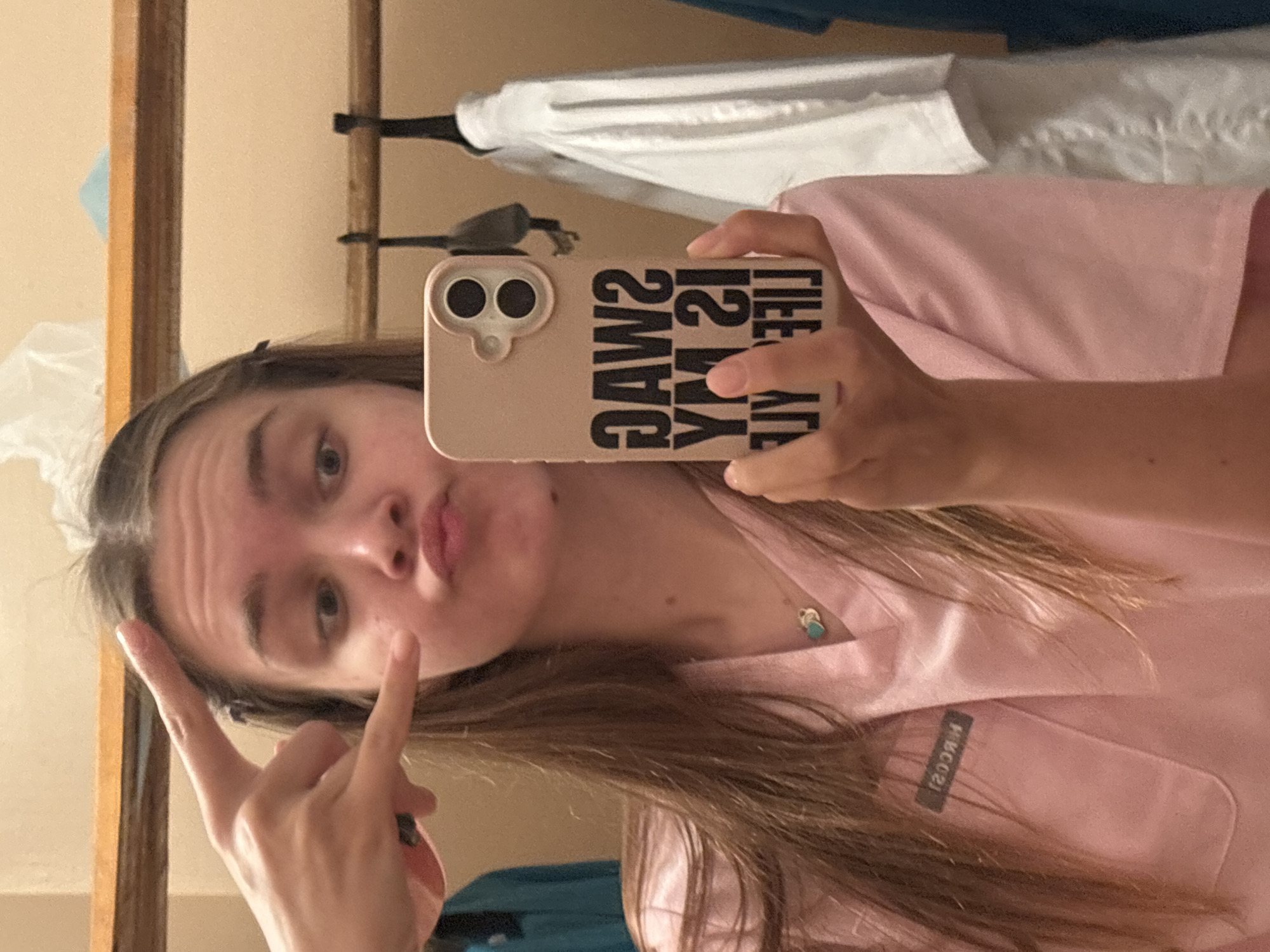 A young woman with long brown hair is taking a selfie in a mirror. She is wearing a pink scrub top with a small black tag on the left chest that reads "NKCOS" in reverse. She is holding her phone in her right hand, which has a pink case with black text that reads "SWAG IS MY LIFE" in reverse. Her left hand is raised with two fingers extended in a peace sign. Her mouth is pursed. In the background, several articles of clothing, including white scrubs, are hanging on a rack. A wooden shelf is visible above the hanging clothes. The environment appears to be a locker room or changing area.